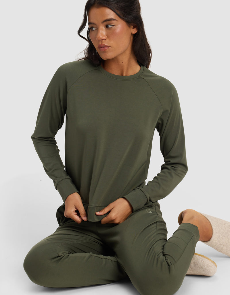 A woman sits on the floor with one knee bent, looking to the side, wearing Cozy Earth’s Women’s Brushed Bamboo Pullover Crew in olive green against a plain light background.