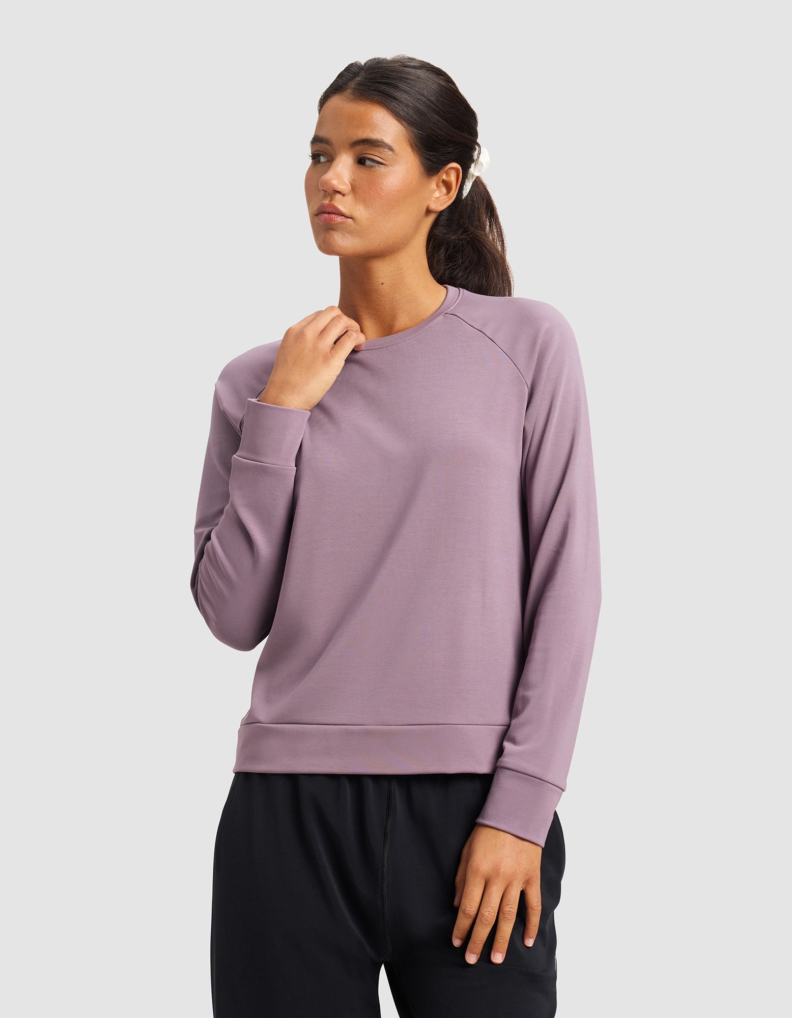 A woman wearing the Cozy Earth Women's Brushed Bamboo Pullover Crew in lavender and black pants stands against a light gray background, looking to the side with one hand touching her collar.