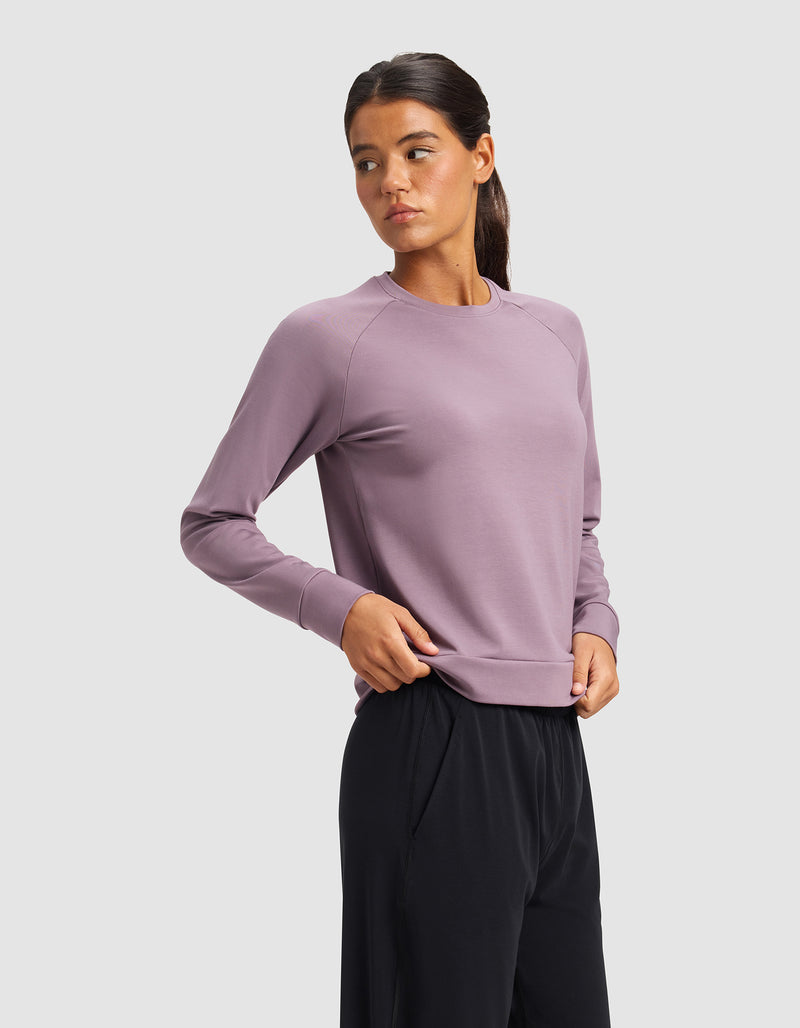 A woman with dark hair in a ponytail wears the Cozy Earth Women's Brushed Bamboo Pullover Crew in mauve and black pants, standing against a light gray background and looking to the side with a neutral expression.
