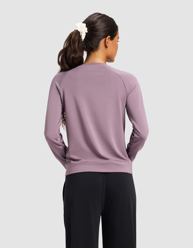 A woman with long brown hair in a white scrunchie is seen from the back, wearing Cozy Earth's Women's Brushed Bamboo Pullover Crew in mauve with black pants, standing against a plain light gray background.