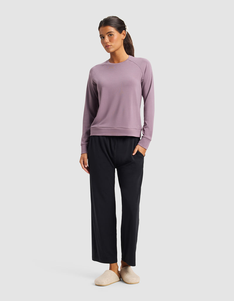 A woman stands against a plain background wearing Cozy Earth's Women's Brushed Bamboo Pullover Crew in lavender, loose black pants, and beige slippers. Her hands are in her pockets and her dark hair is tied back.