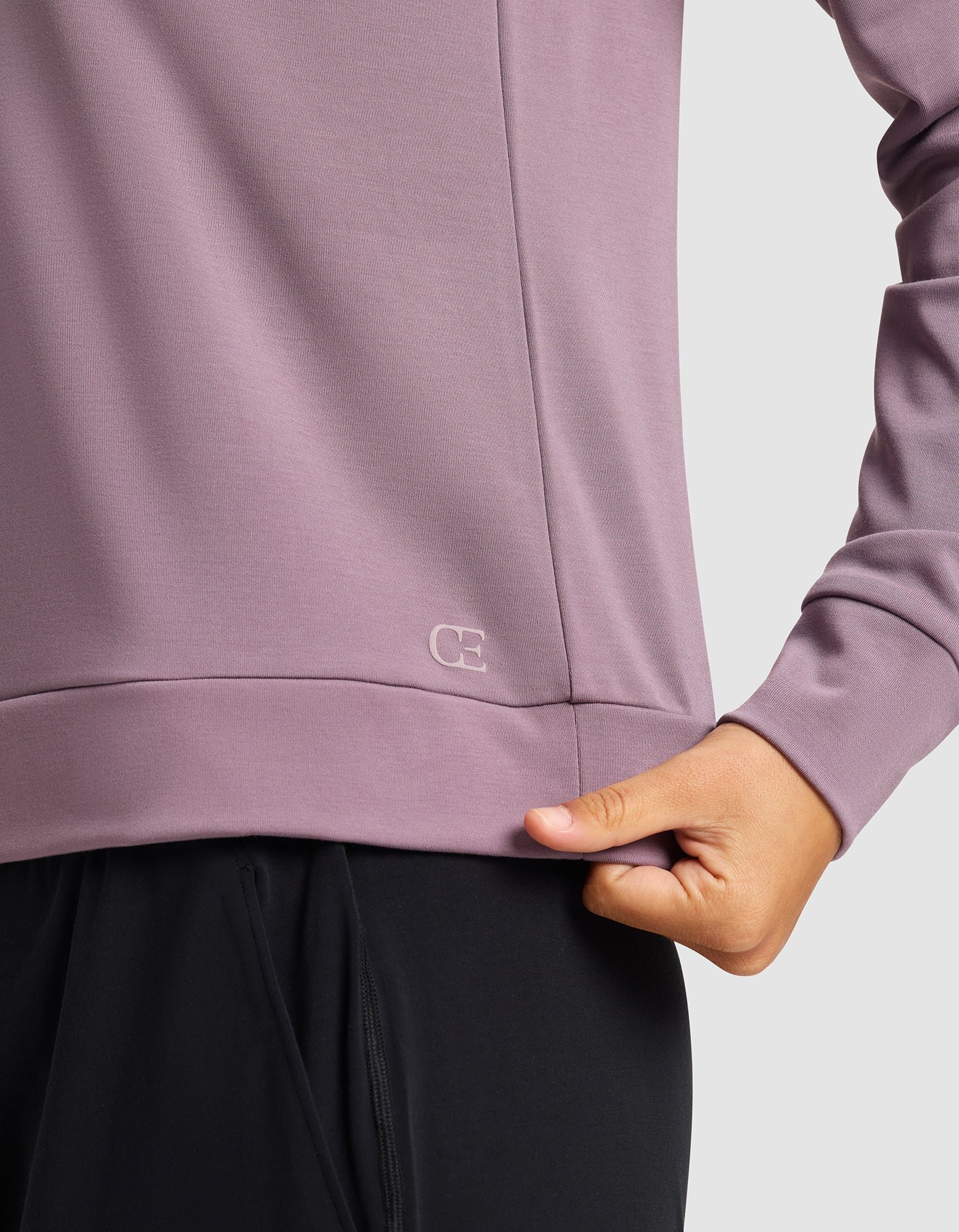 A woman wears the Cozy Earth Women's Brushed Bamboo Pullover Crew, a long-sleeved mauve top featuring a small "CE" logo near the hem and gently pulls its edge; part of her black pants is also visible.