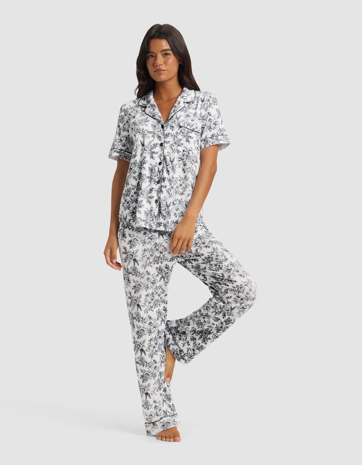 A woman models the Cozy Earth Women's Bamboo Stretch-Knit Short Sleeve & Pant Pajama Set in blue and white floral, standing barefoot against a plain light background.|Color:Jet Black Toile