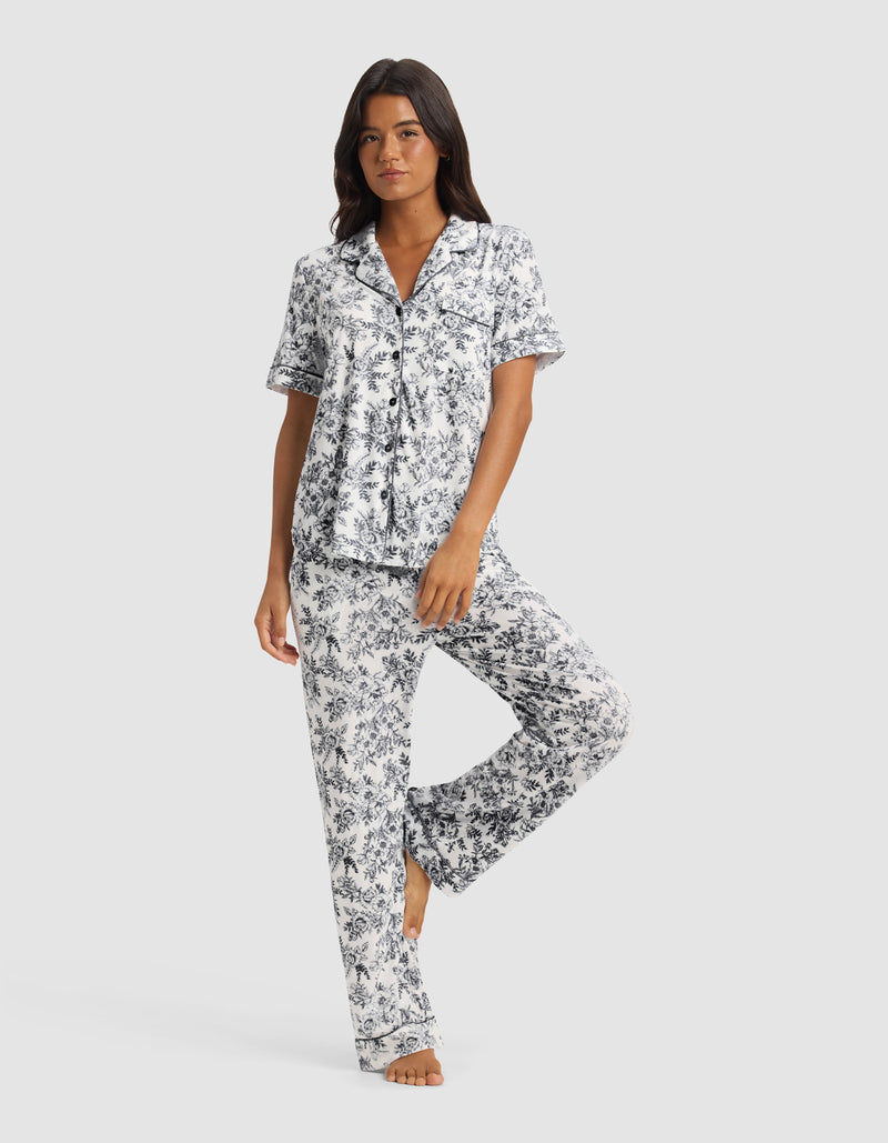 A woman models the Cozy Earth Women's Bamboo Stretch-Knit Short Sleeve & Pant Pajama Set in blue and white floral, standing barefoot against a plain light background.