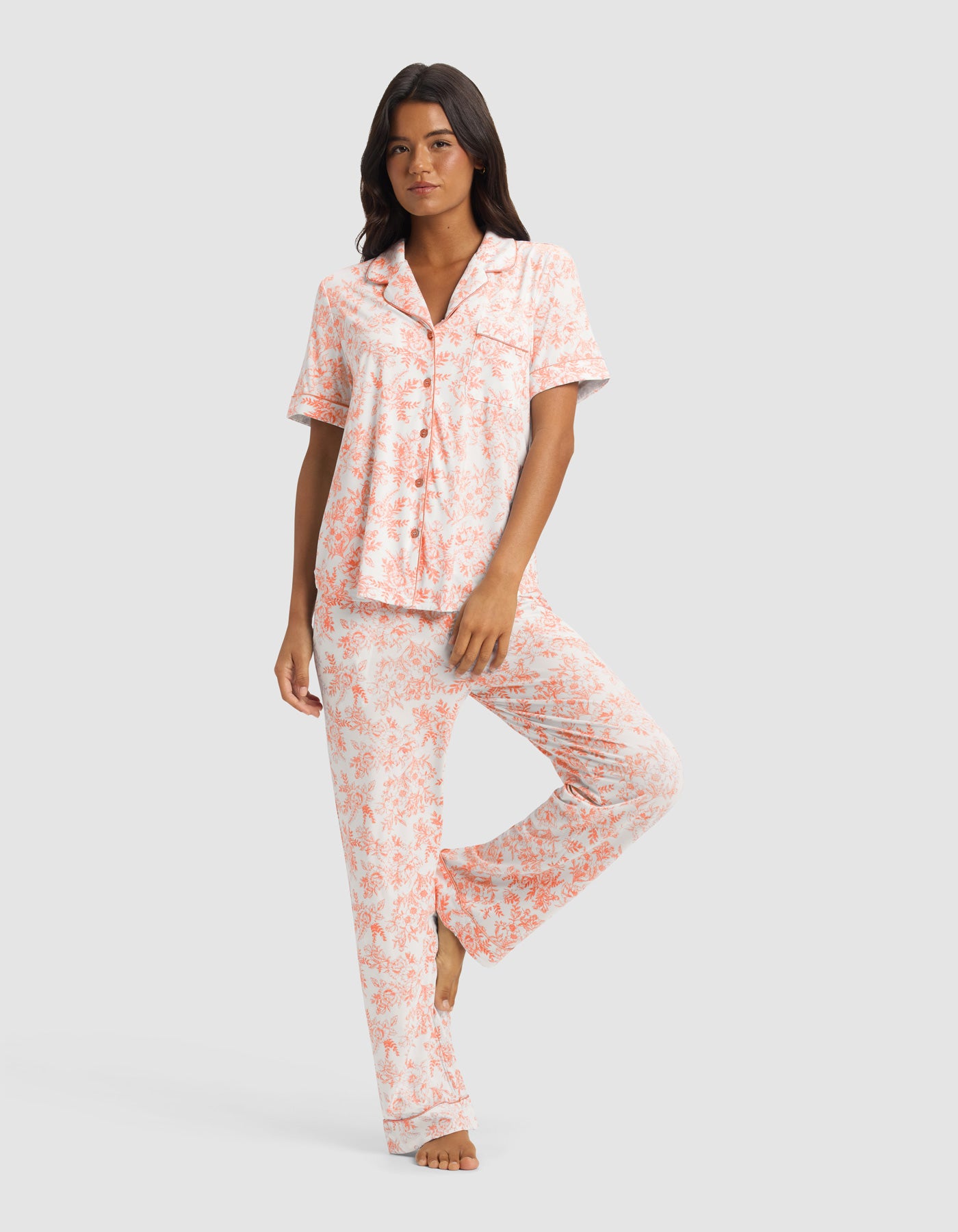 A woman stands barefoot against a plain light background, wearing the Cozy Earth Women's Bamboo Stretch-Knit Short Sleeve & Pant Pajama Set in a light pink and white floral print, with one leg raised and hands by her sides.|Color:Melon Toile