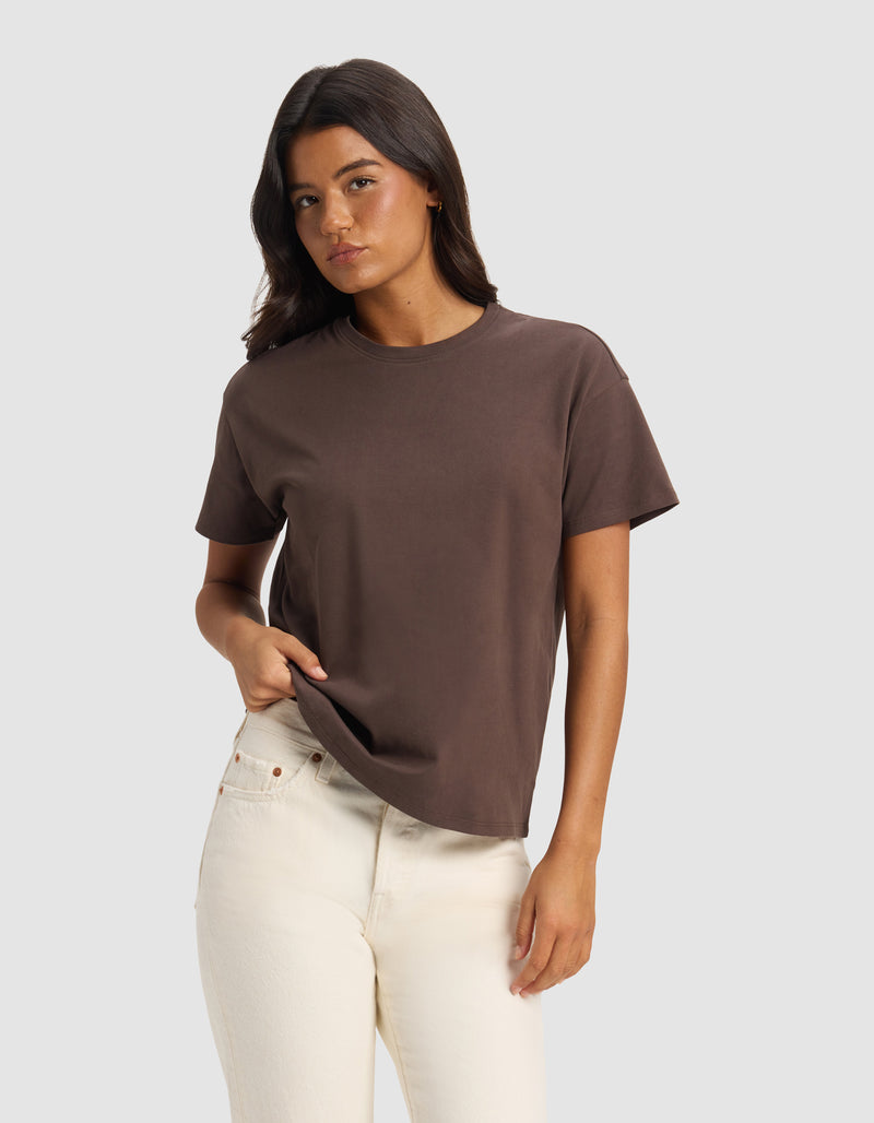 A woman with long dark hair wears the Cozy Earth Women's All Day Tee in brown and off-white jeans, standing against a light gray background with one hand in her pocket and a neutral expression.