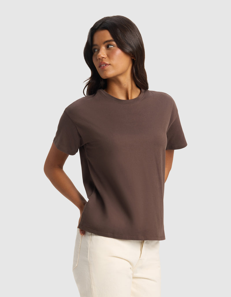 A woman with long dark hair wears Cozy Earth's Women's All Day Tee in brown and off-white pants, standing against a light gray background with her hands behind her back, looking to her left.