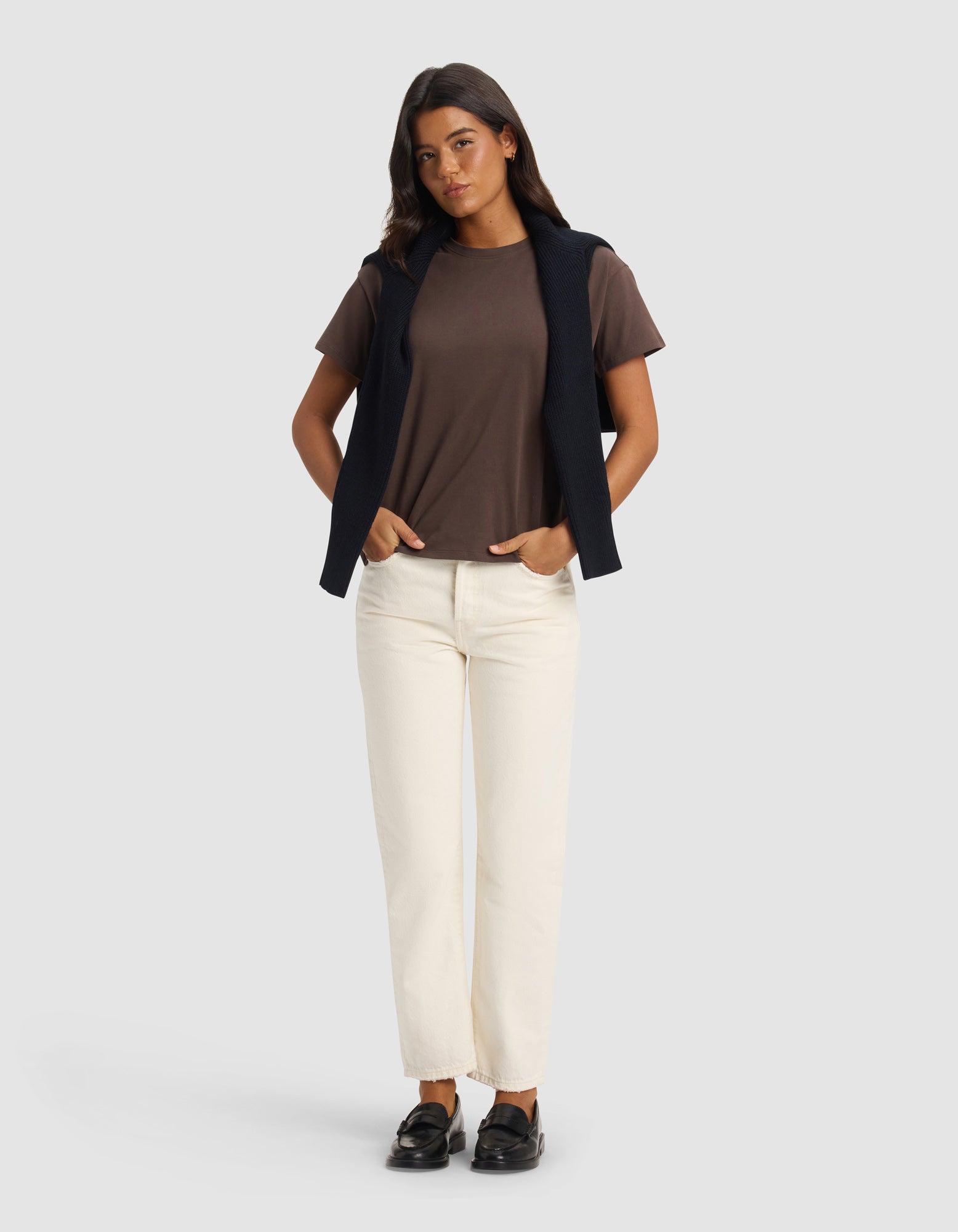 A woman stands against a plain background wearing the Cozy Earth Women's All Day Tee in brown, paired with cream pants, black loafers, and a black sweater draped over her shoulders, with her hands in her pockets.