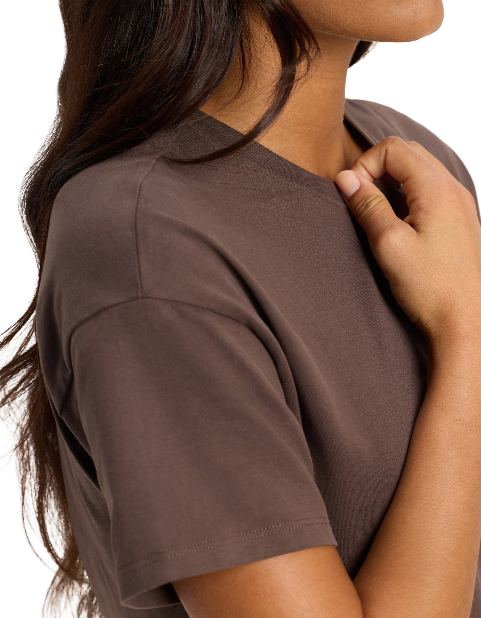 A woman with long brown hair wears the Cozy Earth Women's All Day Tee in brown, holding the collar with her right hand. The image shows her upper torso, arm, and part of her face against a white background.|Color:Cacao