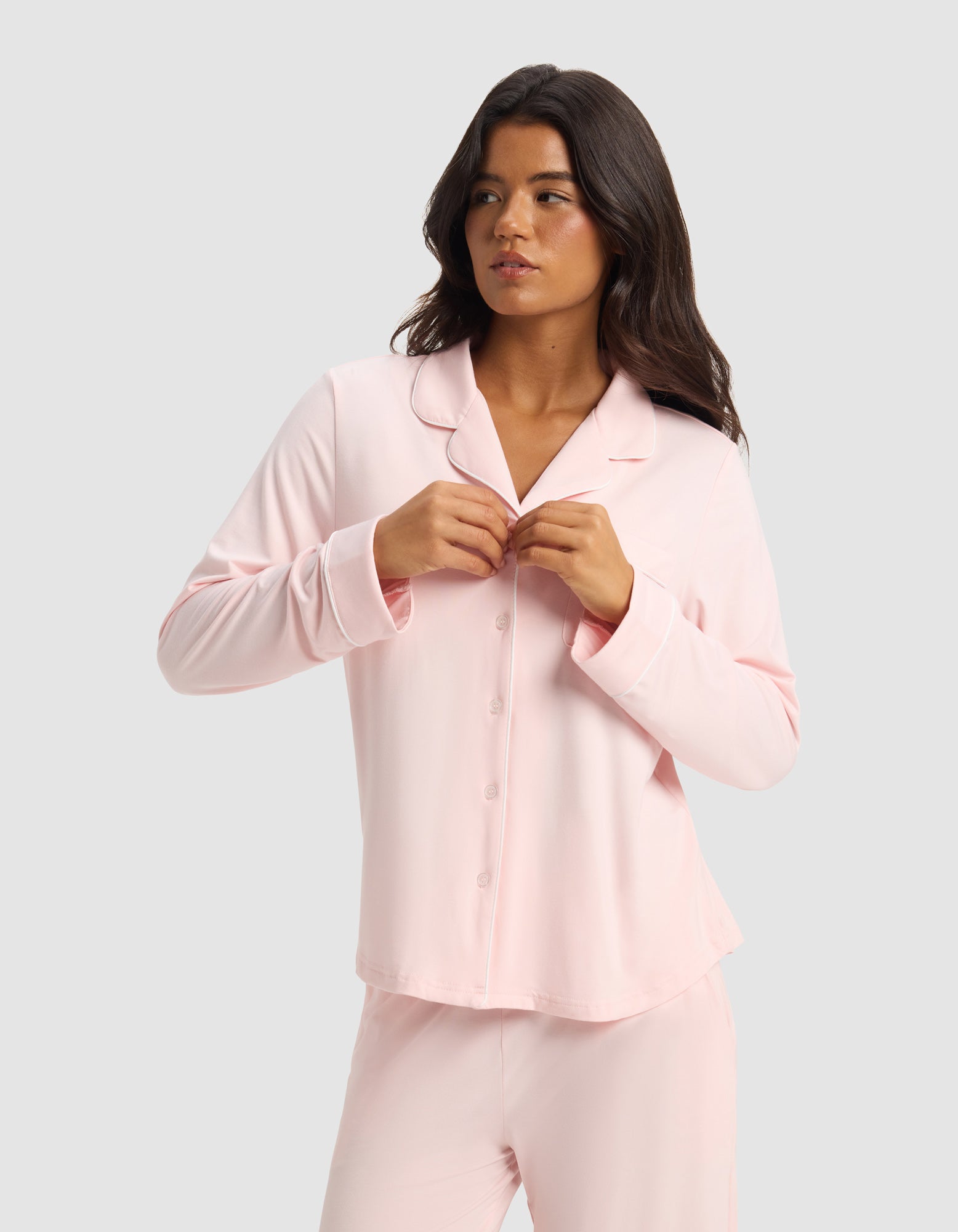 A woman with long dark hair wears the Cozy Earth Women's Bamboo Stretch-Knit Long Sleeve Pajama Set in light pink, fastening a button on her top as she stands against a plain light gray background, looking slightly to her left.
