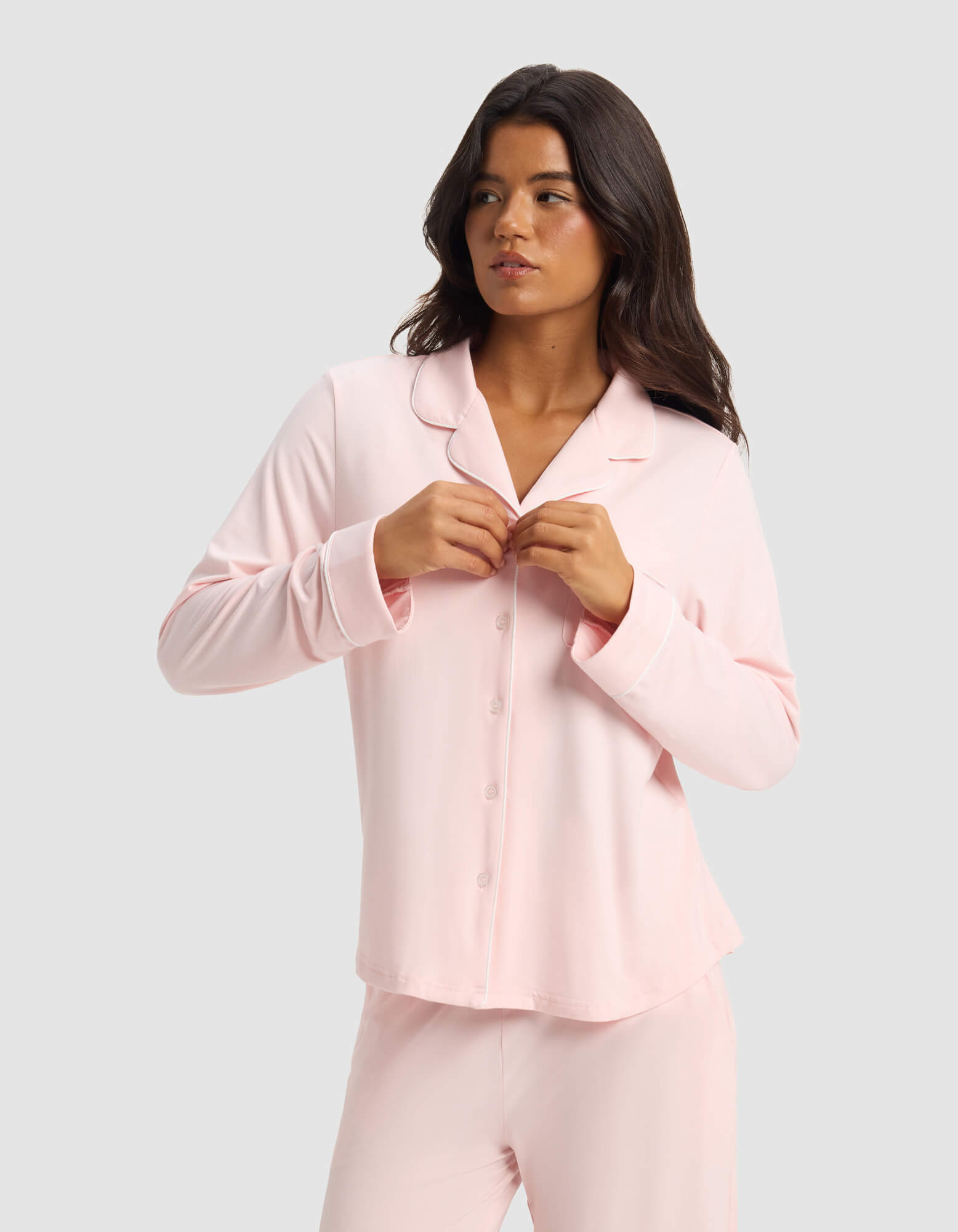 A woman with long dark hair wears the Cozy Earth Women's Bamboo Stretch-Knit Long Sleeve Pajama Set in light pink, fastening a button on her top as she stands against a plain light gray background, looking slightly to her left.|Color:Blossom