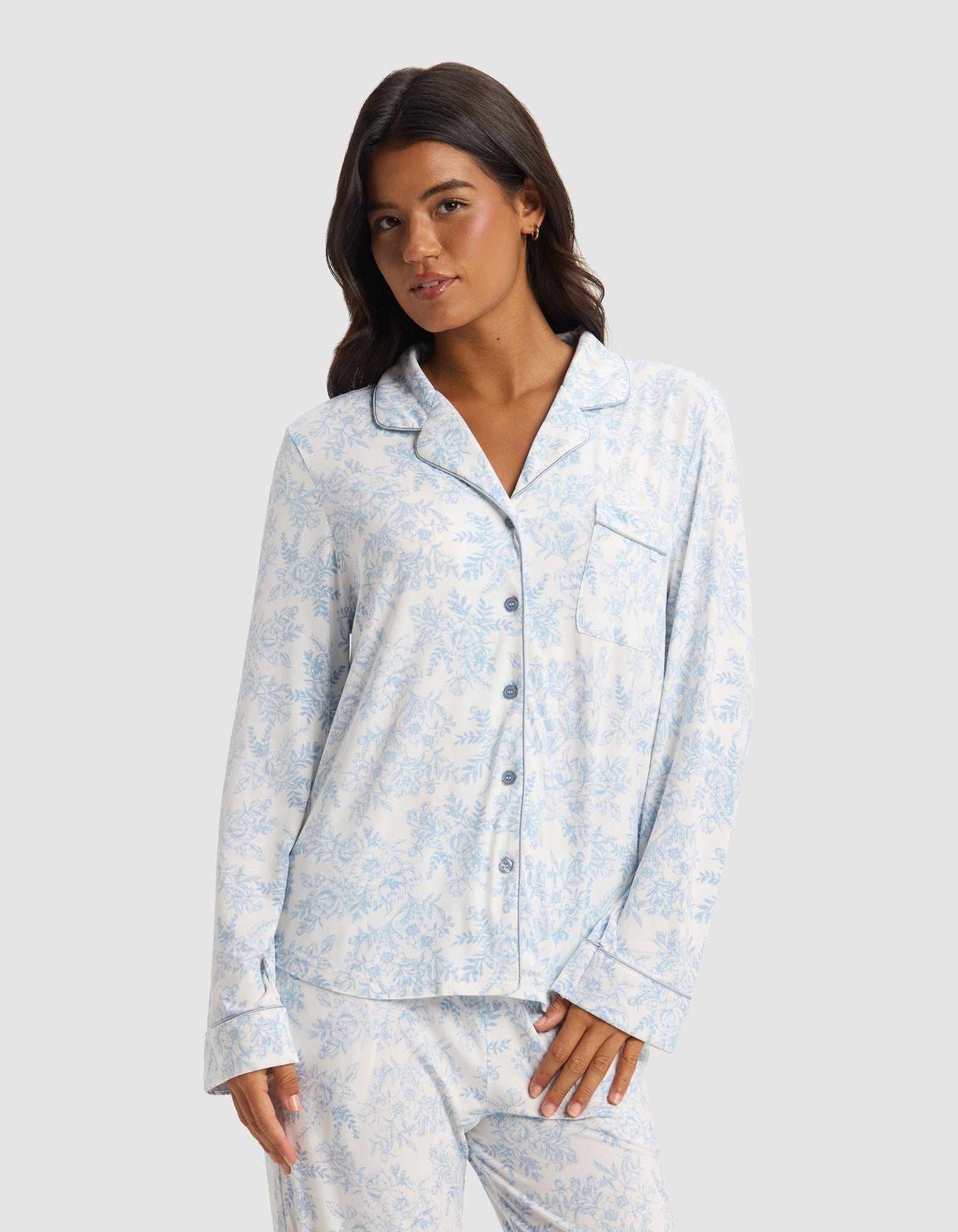 A woman wears the Cozy Earth Women's Bamboo Stretch-Knit Long Sleeve Pajama Set in light blue and white print, featuring a button-up top and matching pants, standing against a plain light gray background.