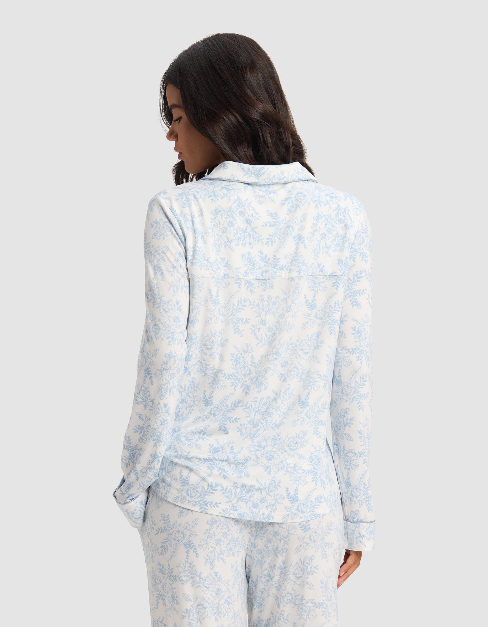 A woman with wavy dark hair is seen from behind, wearing the Cozy Earth Women's Bamboo Stretch-Knit Long Sleeve Pajama Set in a light blue and white floral pattern, standing against a plain light gray background.