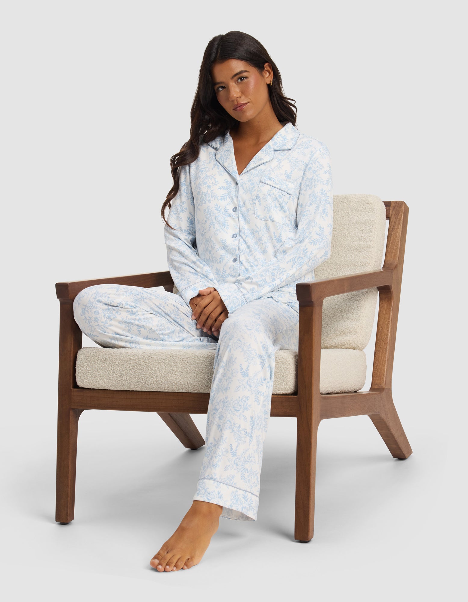 A woman with long dark hair sits cross-legged in a wooden armchair, wearing the Cozy Earth Women's Bamboo Stretch-Knit Long Sleeve Pajama Top, looking relaxed with a neutral expression against a plain background.