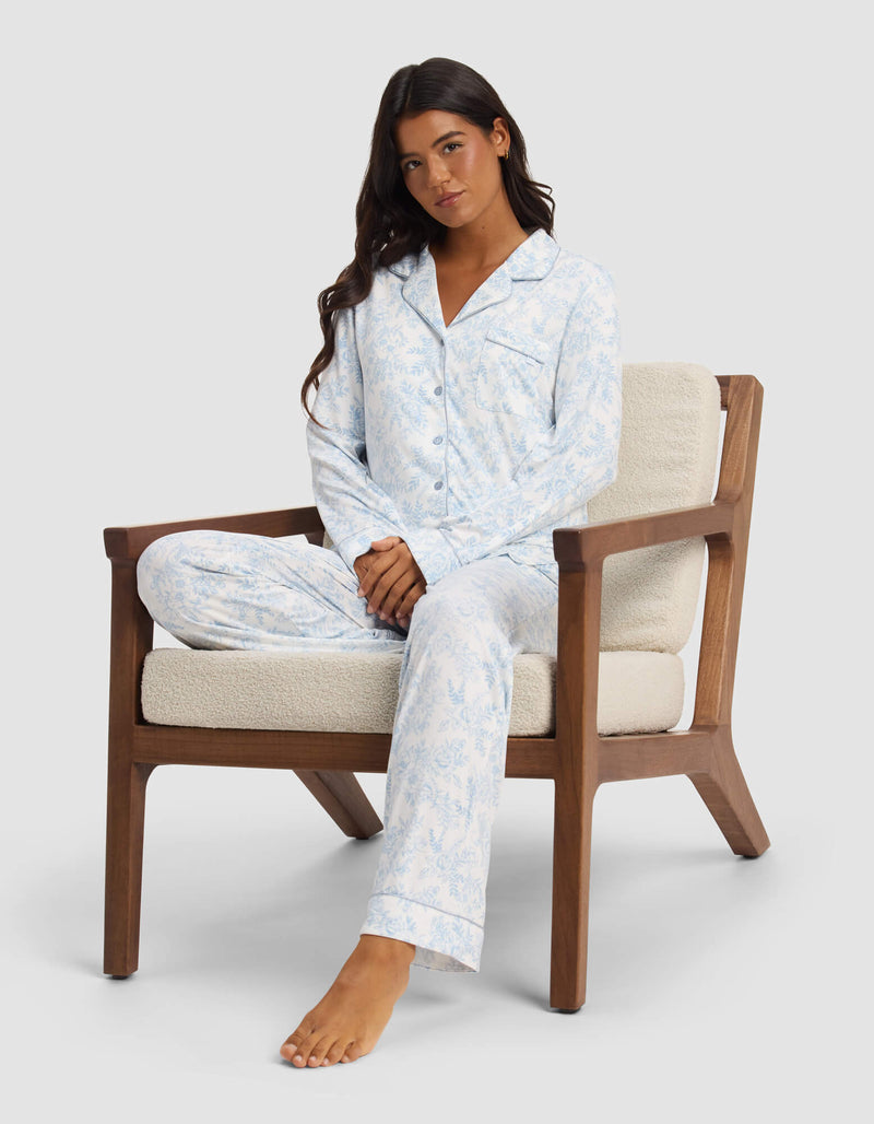 A woman with long dark hair sits cross-legged in a wooden armchair, wearing the Cozy Earth Women's Bamboo Stretch-Knit Long Sleeve Pajama Top, looking relaxed with a neutral expression against a plain background.