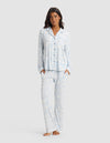 A woman stands barefoot, hands in her pockets, wearing the Cozy Earth Women's Bamboo Stretch-Knit Long Sleeve Pajama Set in a light blue and white pattern, against a plain light gray background.