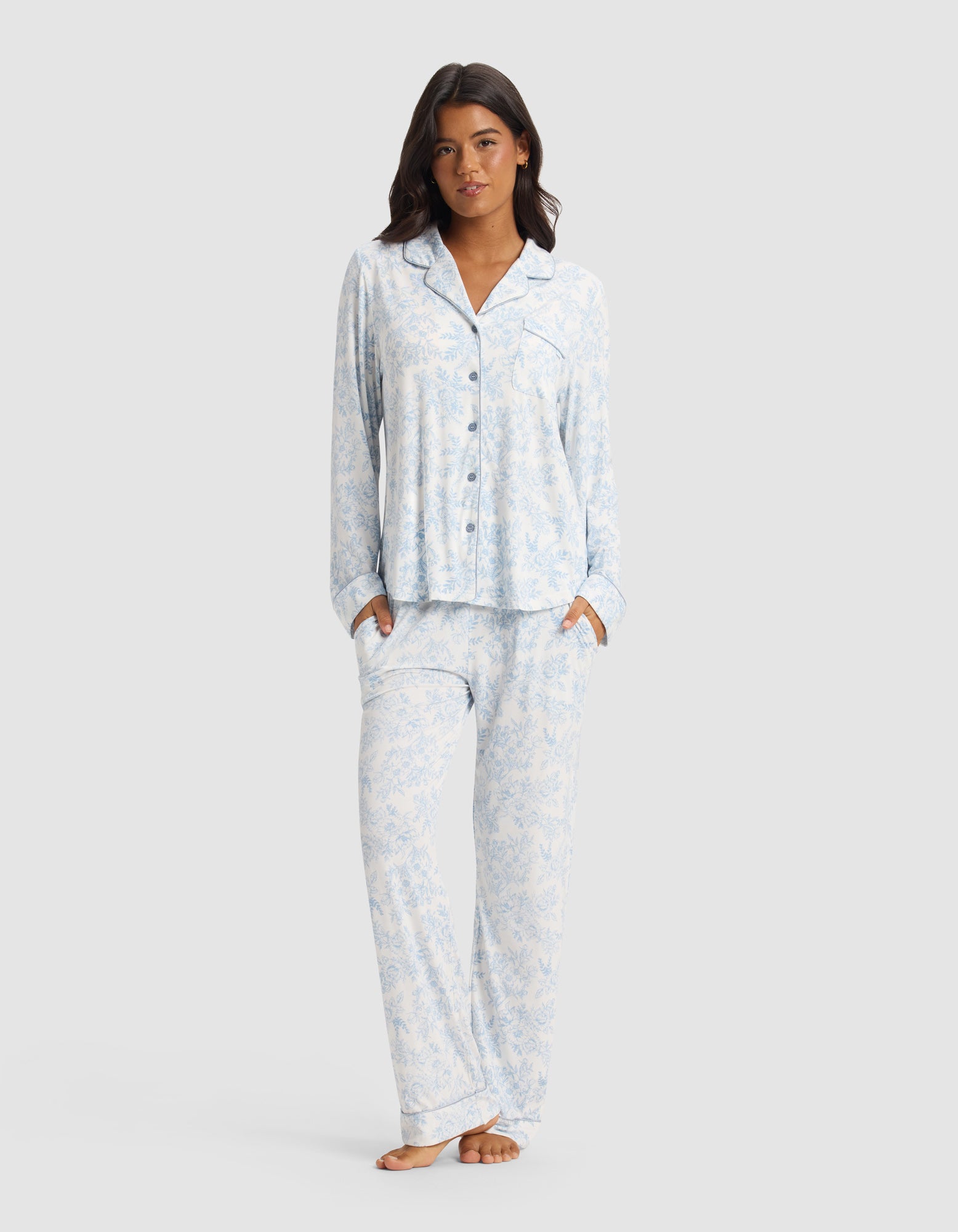 A woman stands barefoot, hands in her pockets, wearing the Cozy Earth Women's Bamboo Stretch-Knit Long Sleeve Pajama Set in a light blue and white pattern, against a plain light gray background.