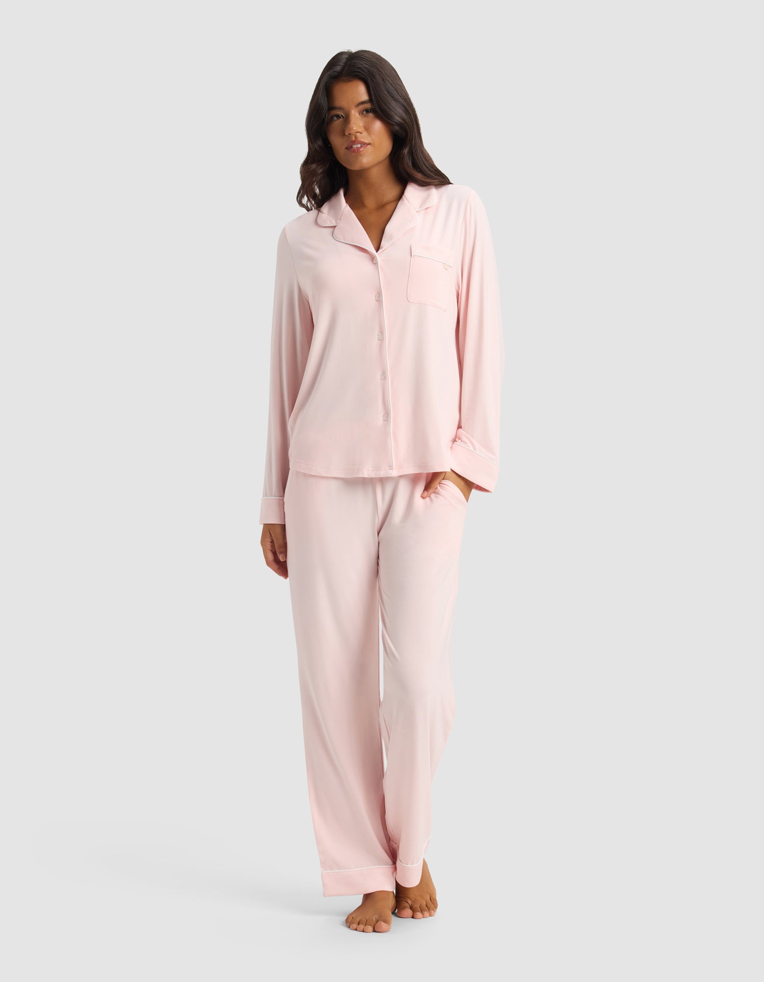 A woman stands barefoot in a Cozy Earth Women’s Bamboo Stretch-Knit Long Sleeve Pajama Set—light pink, button-up top with chest pocket and matching loose pants—against a plain light gray background.