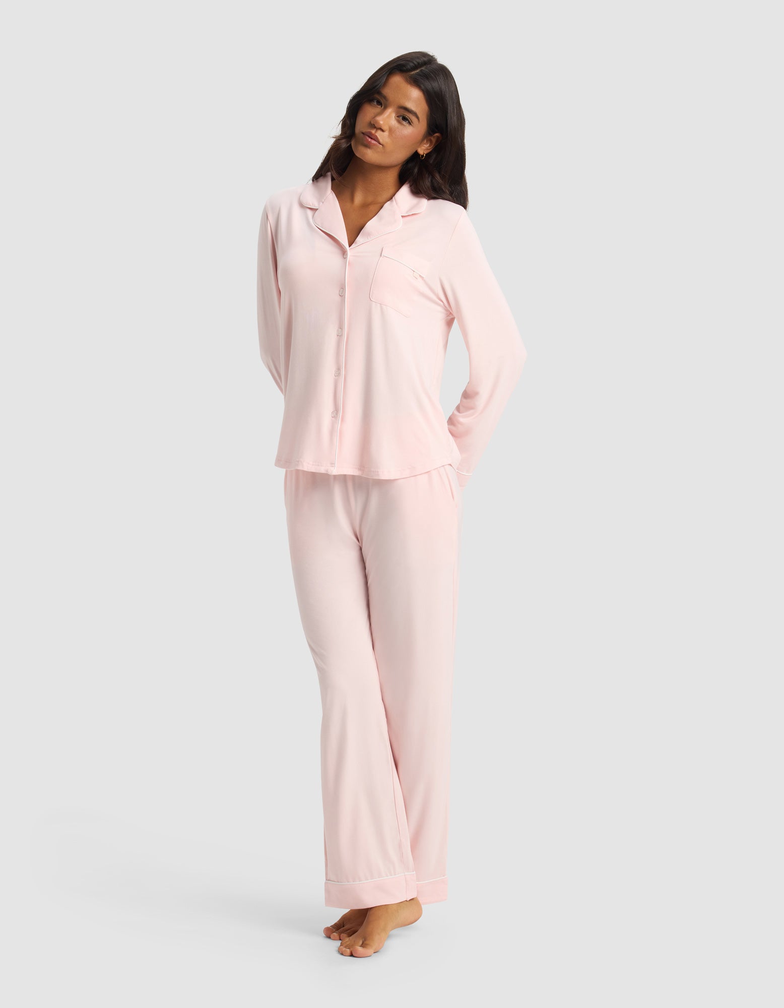 A woman with long dark hair stands barefoot against a light gray background, wearing Cozy Earth’s Women's Bamboo Stretch-Knit Long Sleeve Pajama Set—light pink, with a button-up top and matching pants. Her arms are relaxed behind her back.