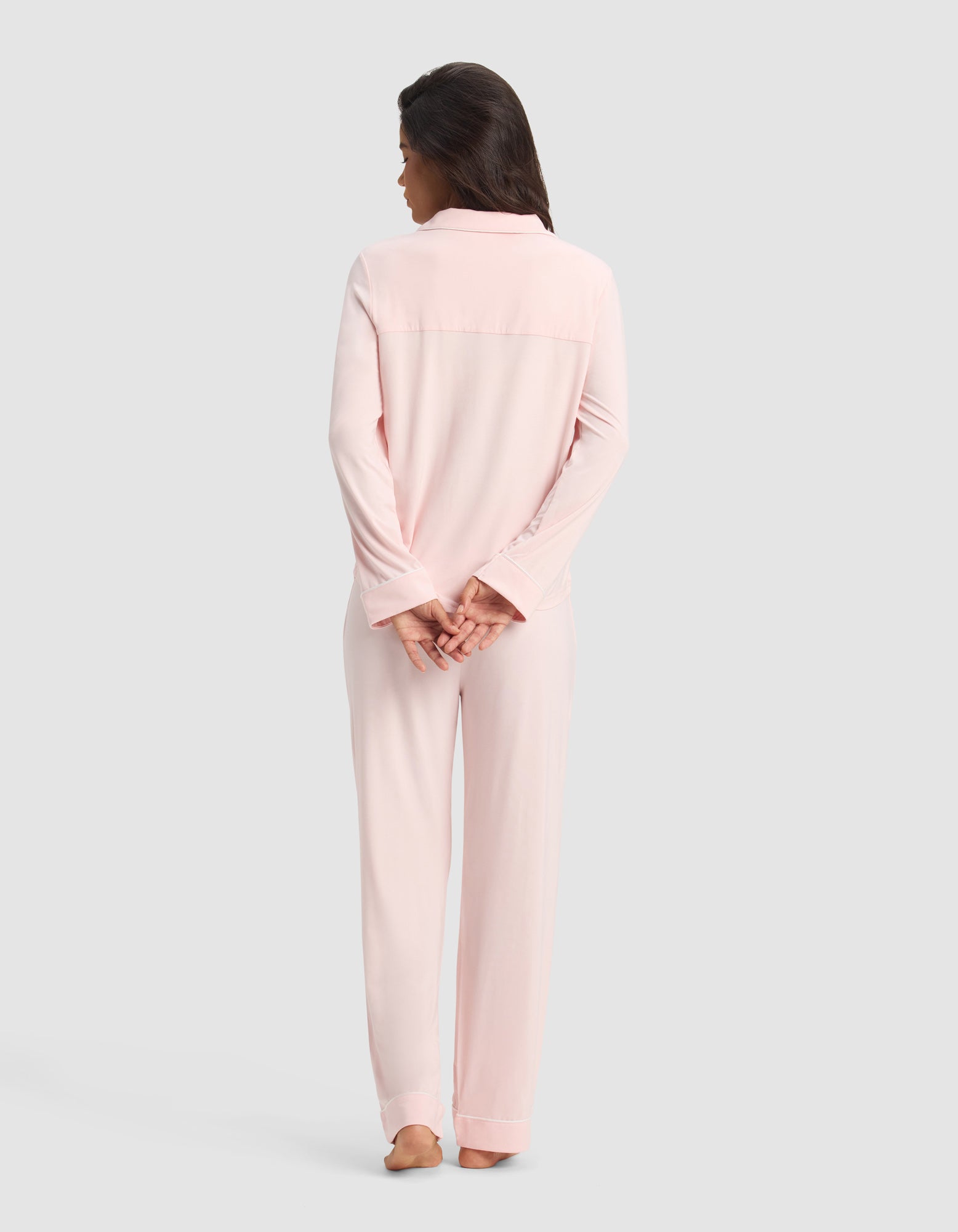 A woman stands barefoot facing away in the Cozy Earth Women's Bamboo Stretch-Knit Long Sleeve Pajama Set in light pink. Her dark hair is loose, hands clasped behind her back, against a plain light gray background.