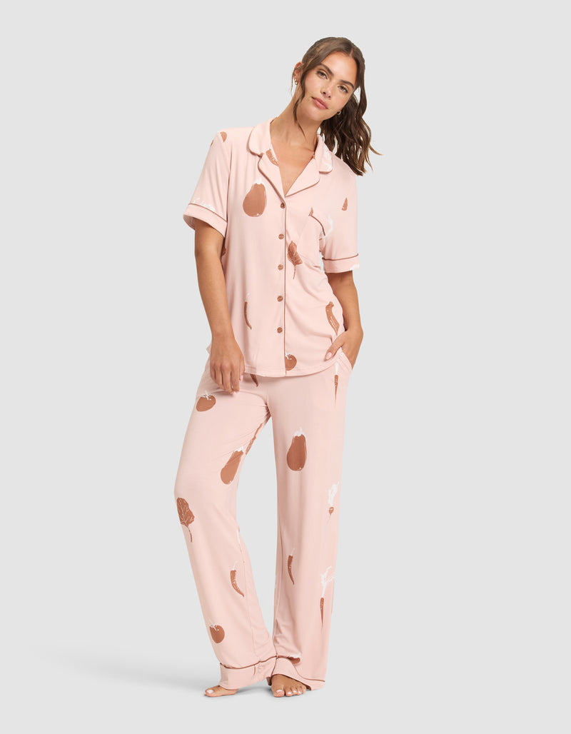 A woman stands barefoot wearing Cozy Earth's Women's Bamboo Stretch-Knit Short Sleeve & Pant Pajama Set in light pink with an abstract brown and white print. The set features a collared, button-up top and matching pants. She has one hand in her pocket, looking relaxed.