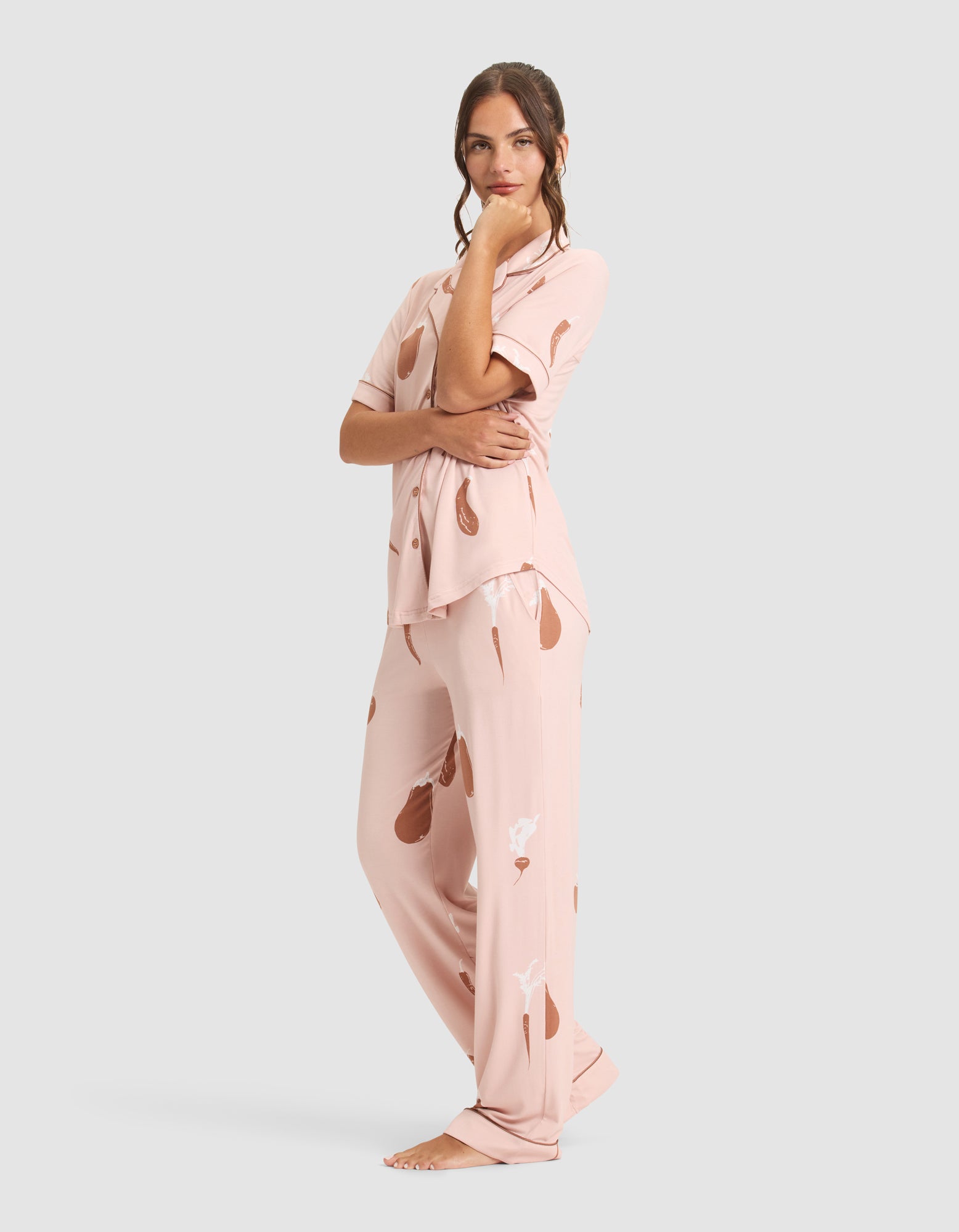 A woman stands barefoot on a gray background, wearing the Cozy Earth Women's Bamboo Stretch-Knit Short Sleeve Pajama Set in light pink with a brown abstract pattern, posing with one hand on her chin and looking at the camera. 