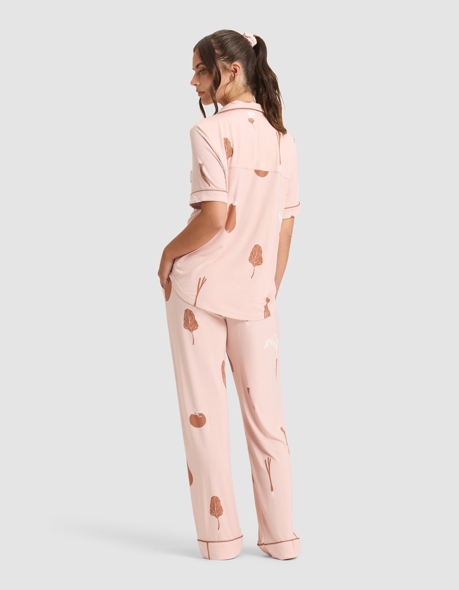 A person faces away in Cozy Earth's Women's Bamboo Stretch-Knit Short Sleeve & Pant Pajama Set—light pink with a brown tree and apple print, featuring a collared short-sleeve top and loose long pants, against a plain gray background. 