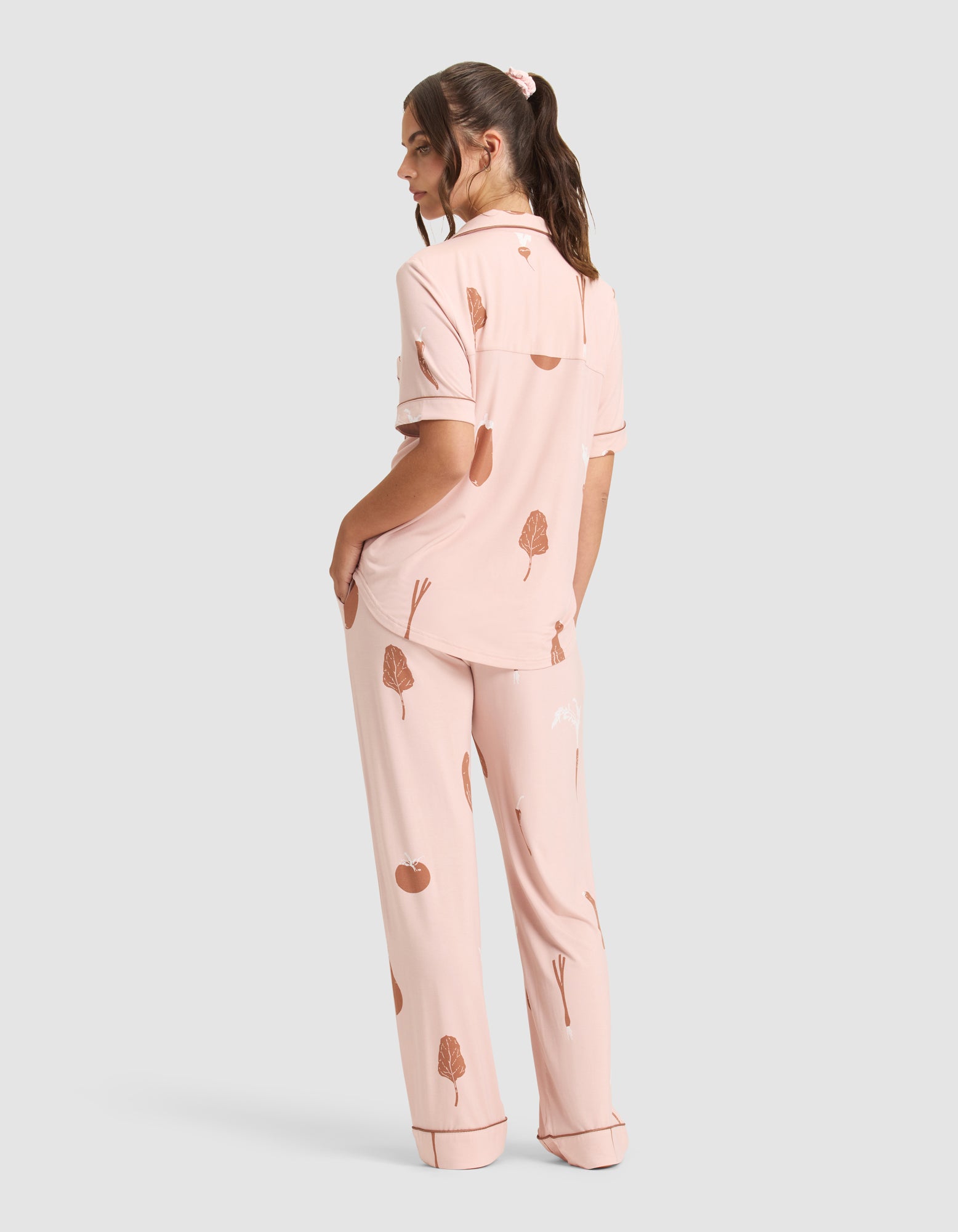 A person faces away in Cozy Earth's Women's Bamboo Stretch-Knit Short Sleeve & Pant Pajama Set—light pink with a brown tree and apple print, featuring a collared short-sleeve top and loose long pants, against a plain gray background. 