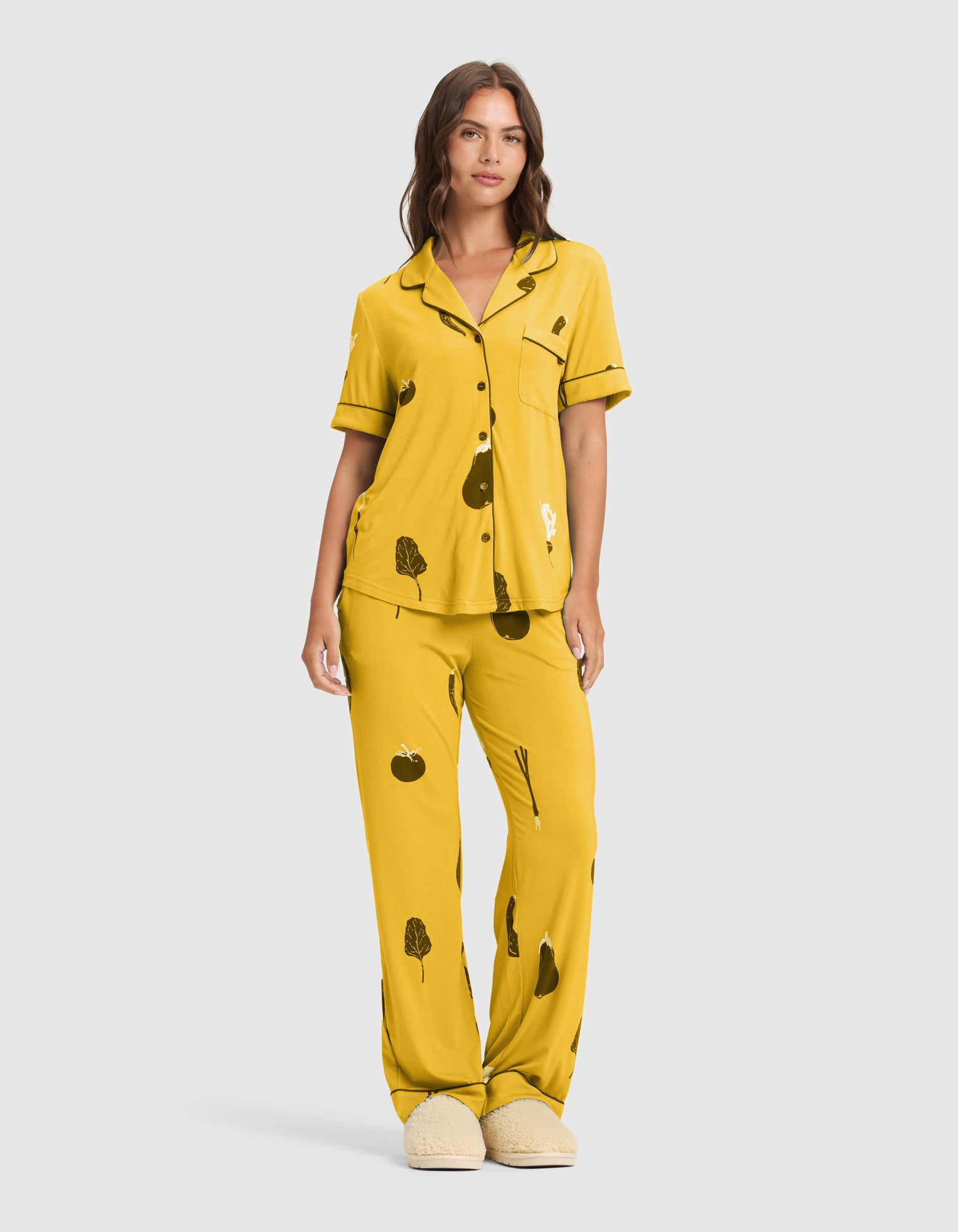 A woman with long brown hair and a neutral expression wears HIDE's Women's Bamboo Stretch-Knit Pajama Pant (Veggie)—a matching yellow set with black veggie-themed patterns, button-up short-sleeve top, long pants, and beige slippers. 