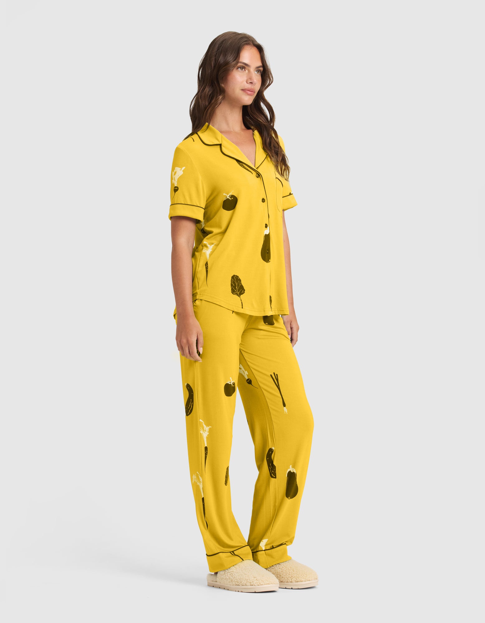 A woman stands facing slightly right, wearing HIDE's Women's Bamboo Stretch-Knit Pajama Pant (Veggie) featuring yellow fabric with black piping and vegetable prints, paired with light slippers, against a plain light gray background. 