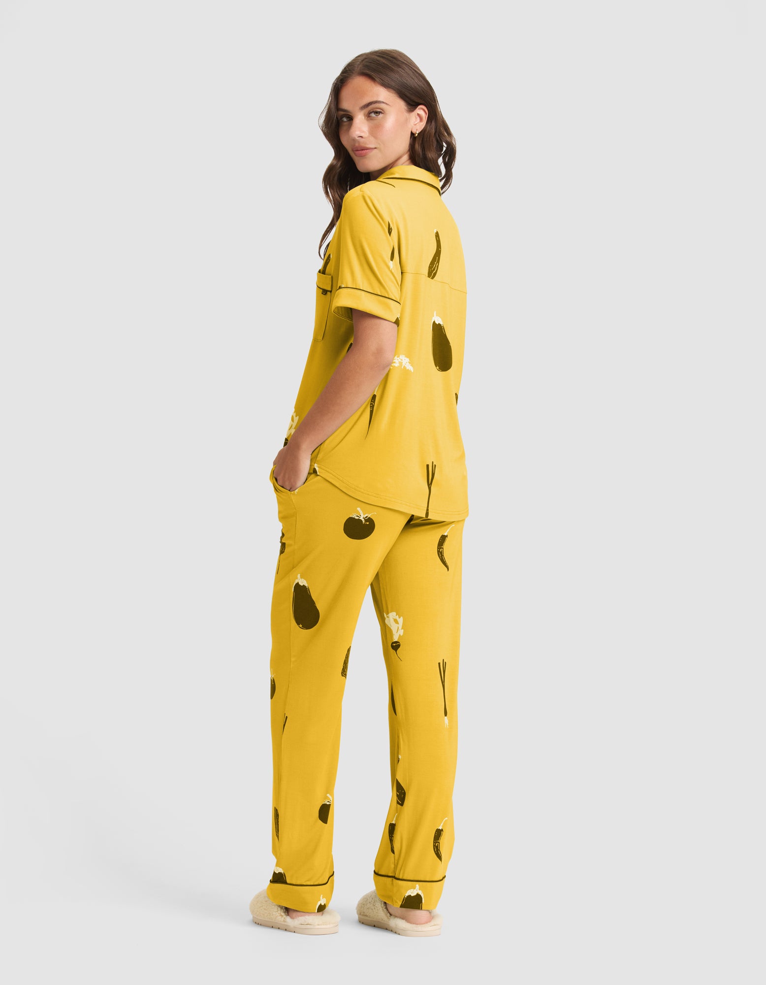 A woman with long brown hair stands against a light gray background, wearing HIDE's Women's Bamboo Stretch-Knit Pajama Pant (Veggie) in yellow with a black vegetable print, facing away from the camera and turning her head to the side. 