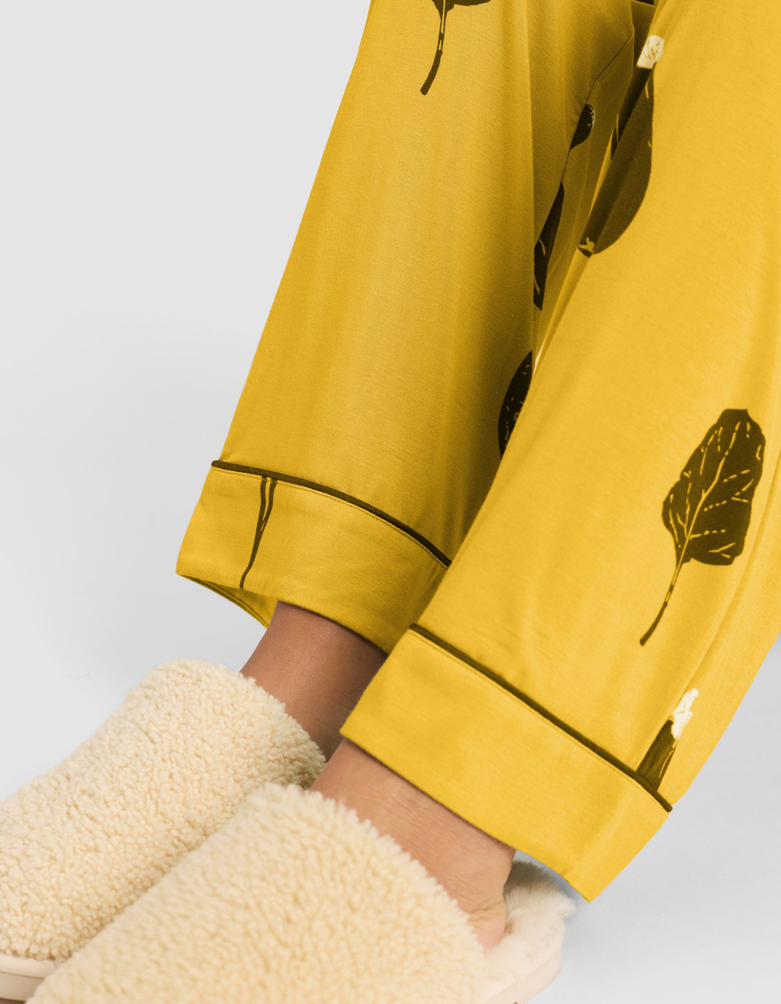 Close-up of a person wearing HIDE Women's Bamboo Stretch-Knit Pajama Pant (Veggie) and cream-colored fuzzy slippers, standing on a light surface. Only their lower legs and feet are visible. 
