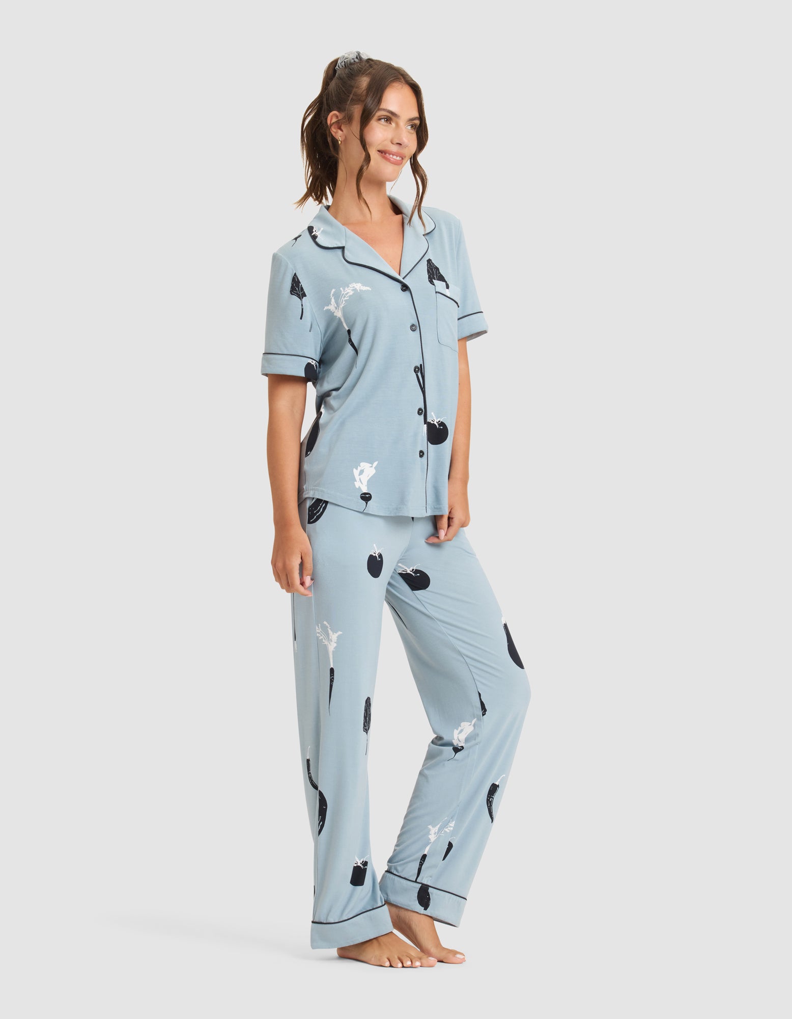 A woman wears the Cozy Earth Women's Bamboo Stretch-Knit Short Sleeve & Pant Pajama Set featuring blue fabric and black-and-white vegetable prints, standing barefoot and smiling against a plain white background. |Color:Veggies in Smokey Blue