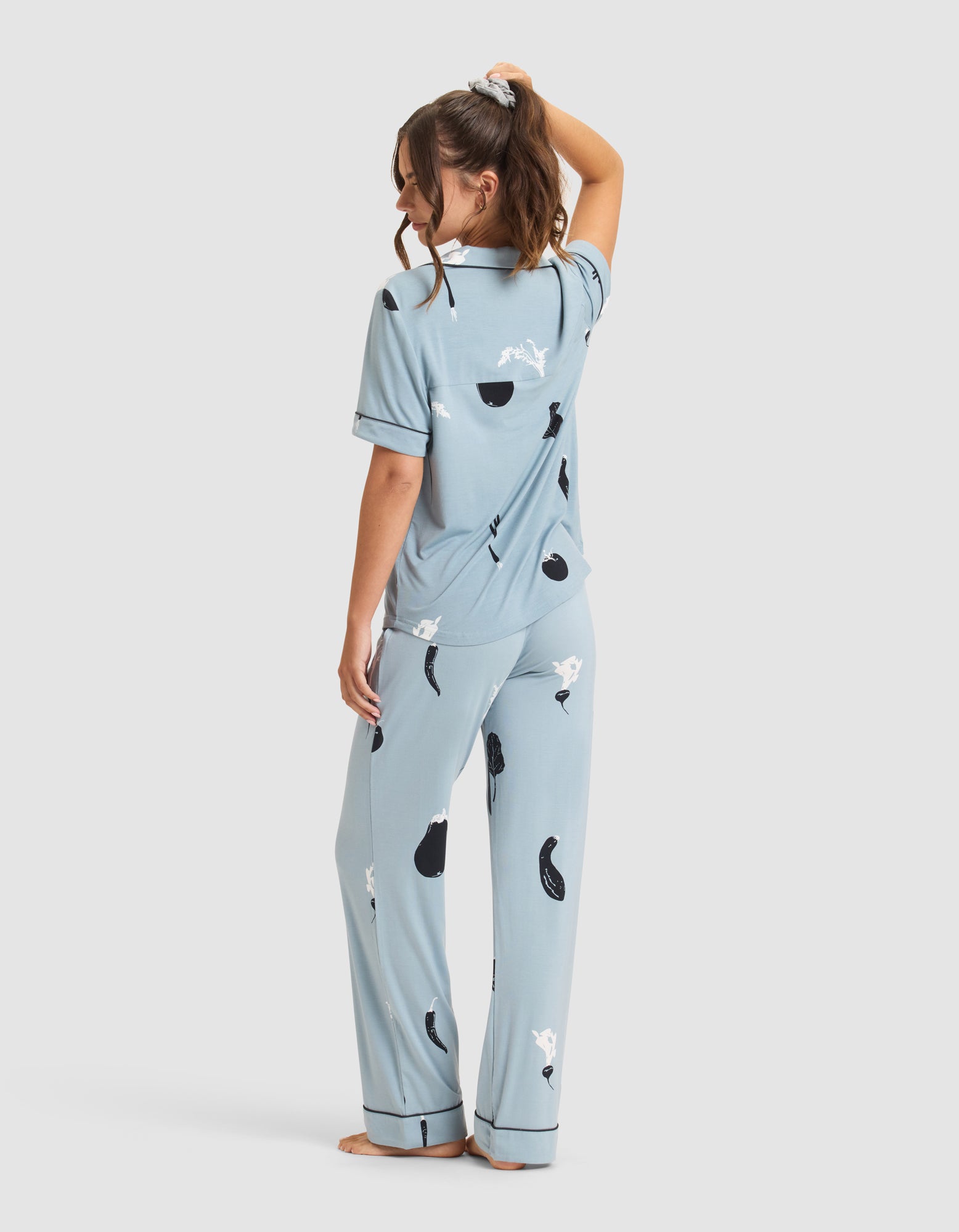 A woman with brown hair in a loose ponytail stands barefoot, glancing over her shoulder while wearing Cozy Earth's Women's Bamboo Stretch-Knit Short Sleeve Pajama Set in a light blue abstract print. 