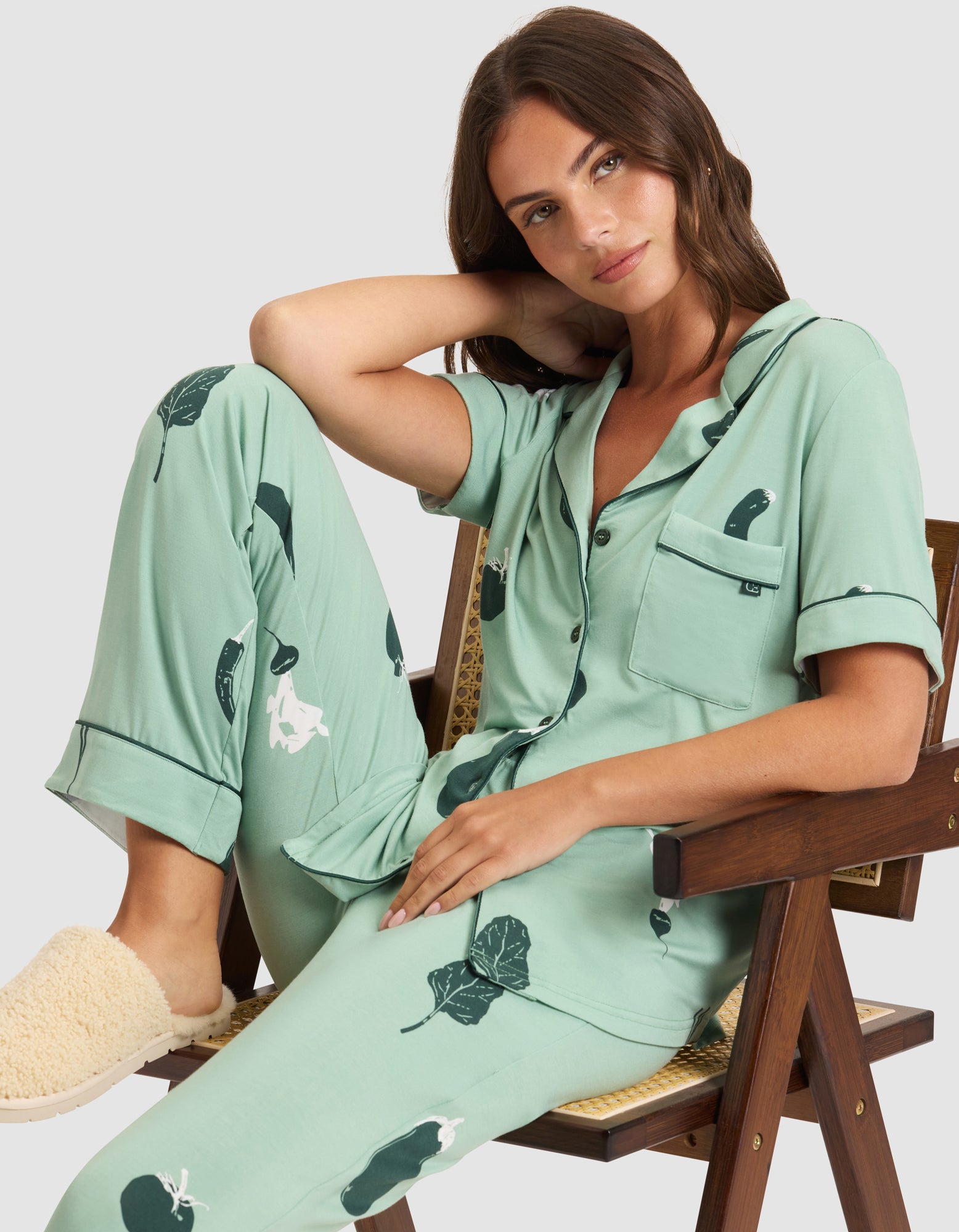 A woman with long brown hair wears the Cozy Earth Women's Bamboo Stretch-Knit Short Sleeve Pajama Set. She sits on a wooden chair, looking at the camera with a relaxed expression. Beige slippers are visible on her feet. 