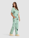 A woman with long brown hair wears HIDE Women's Bamboo Stretch-Knit Pajama Pants in light green with black abstract prints, paired with a matching short-sleeve button-up top and beige slippers, hands in her pockets and gazing to the side. |Color:Veggies in Wintergreen