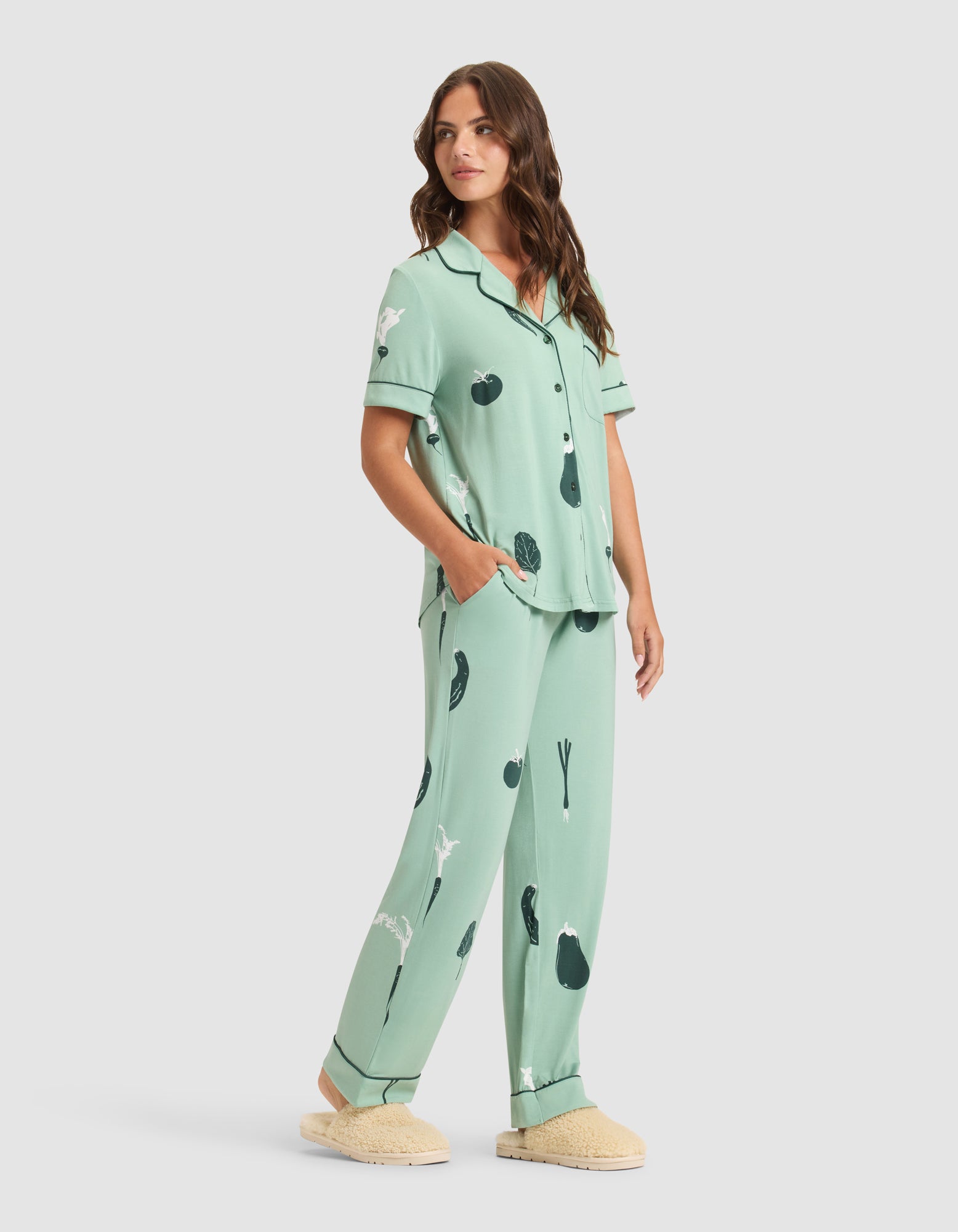 A woman with long brown hair wears HIDE Women's Bamboo Stretch-Knit Pajama Pants in light green with black abstract prints, paired with a matching short-sleeve button-up top and beige slippers, hands in her pockets and gazing to the side. 