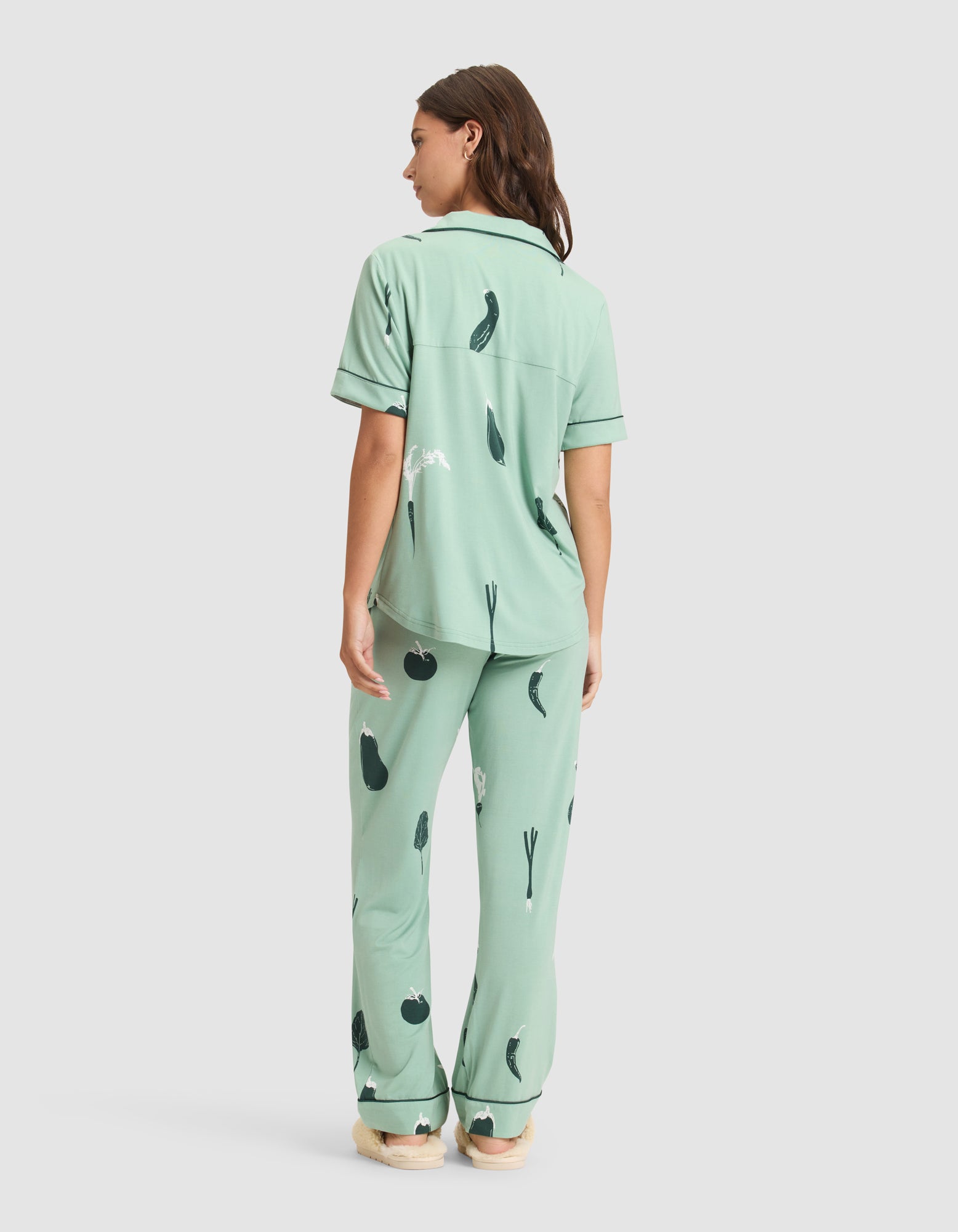 A woman with brown hair stands on a white background wearing Cozy Earth’s Women's Bamboo Stretch-Knit Short Sleeve Pajama Set in mint green with dark green vegetable prints, featuring a button-up top and long pants. 
