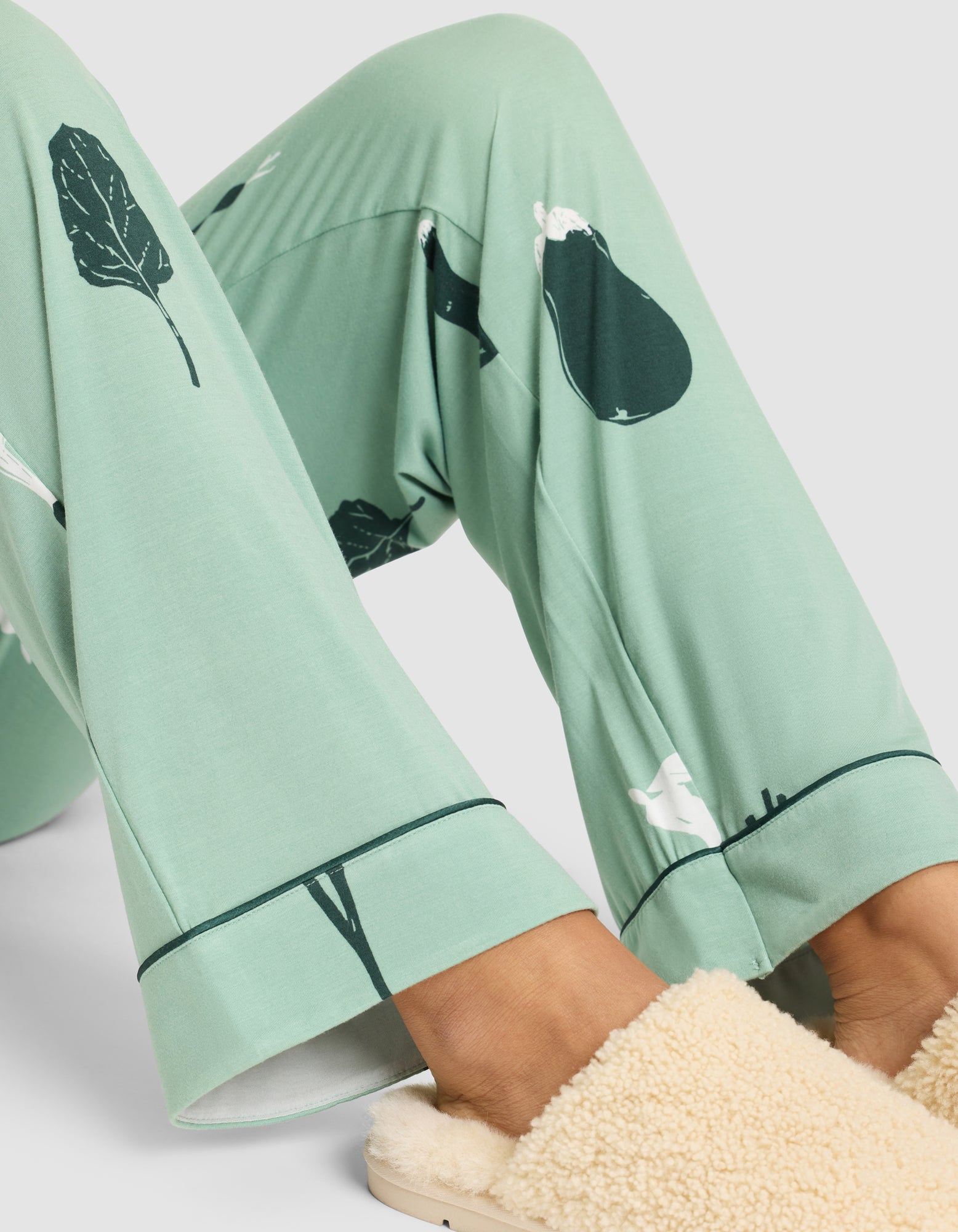 A person sits with bent legs, wearing HIDE Women's Bamboo Stretch-Knit Pajama Pant in light green with dark vegetable prints and beige fluffy slippers. The image highlights their lower legs and feet. 