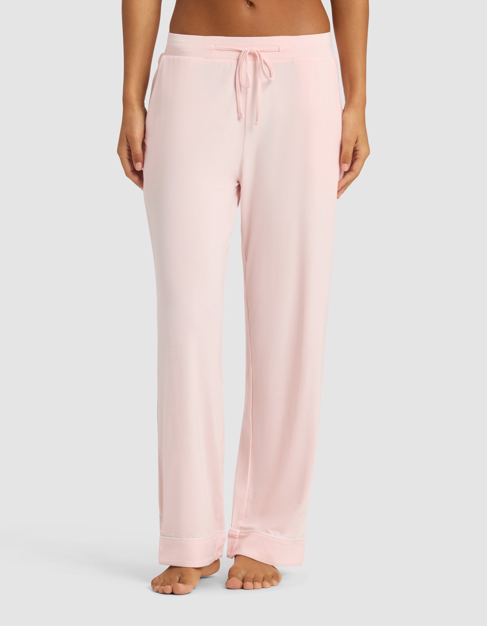 A woman wears Cozy Earth's Women's Bamboo Stretch-Knit Long Sleeve Pajama Set in light pink with a drawstring waist, standing barefoot against a plain light gray background.