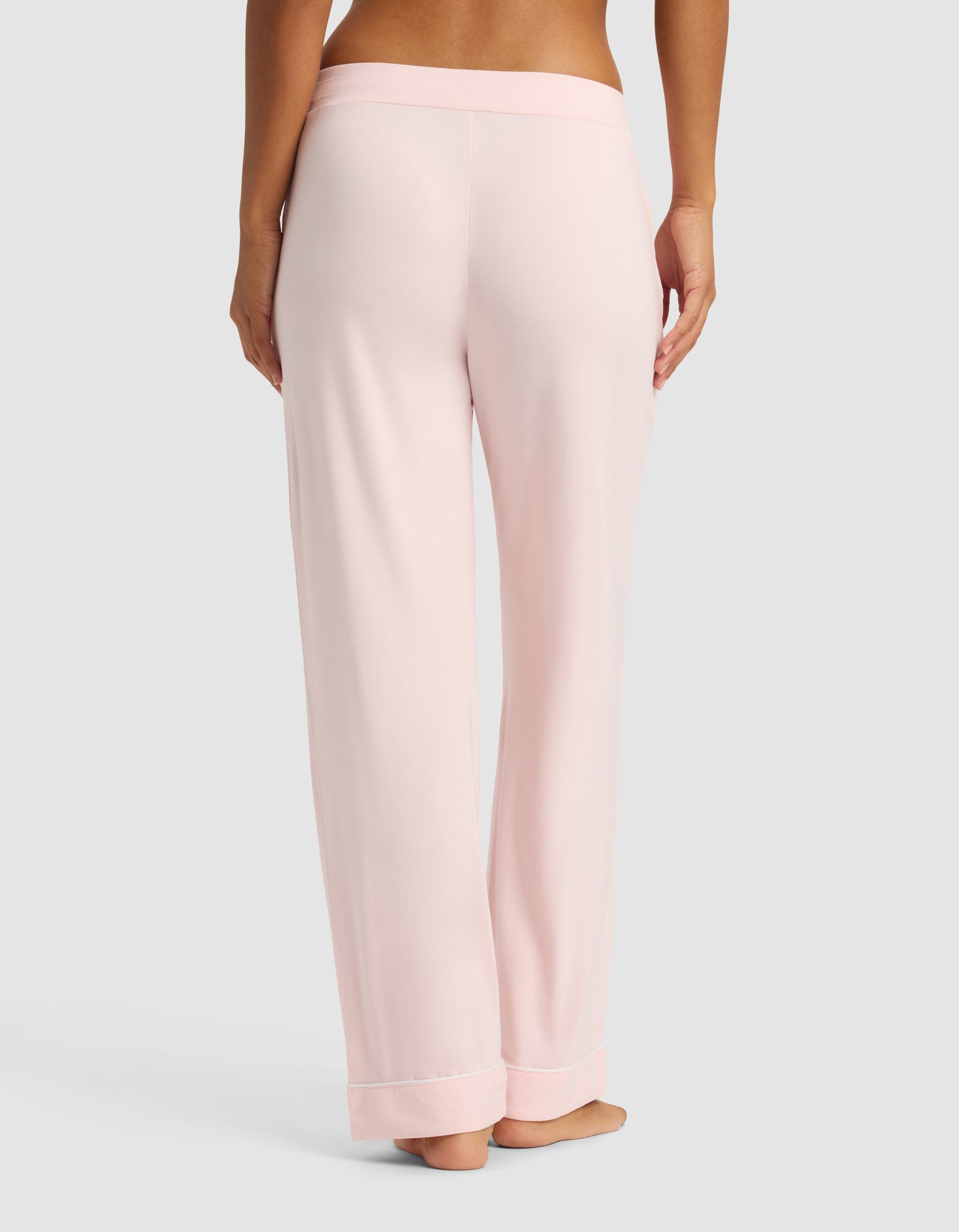 A woman is shown from the waist down wearing Cozy Earth Women's Bamboo Stretch-Knit Long Sleeve Pajama Set pants in light pink, standing barefoot against a plain gray background.