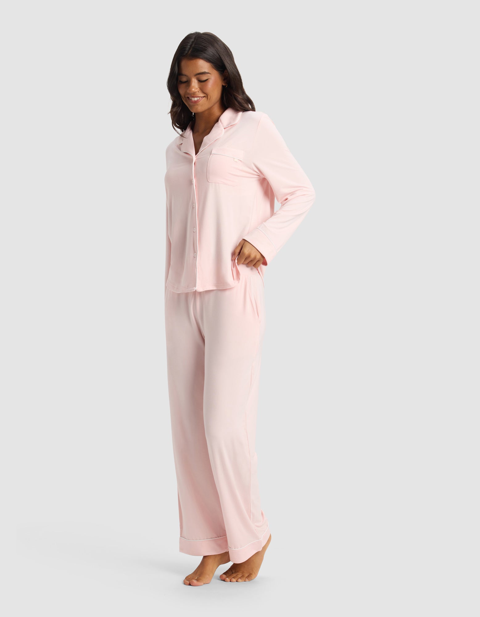 A woman with long dark hair stands barefoot, smiling and wearing Cozy Earth’s Women's Bamboo Stretch-Knit Pajama Pant in light pink, paired with a matching long-sleeved top, against a plain light gray background.