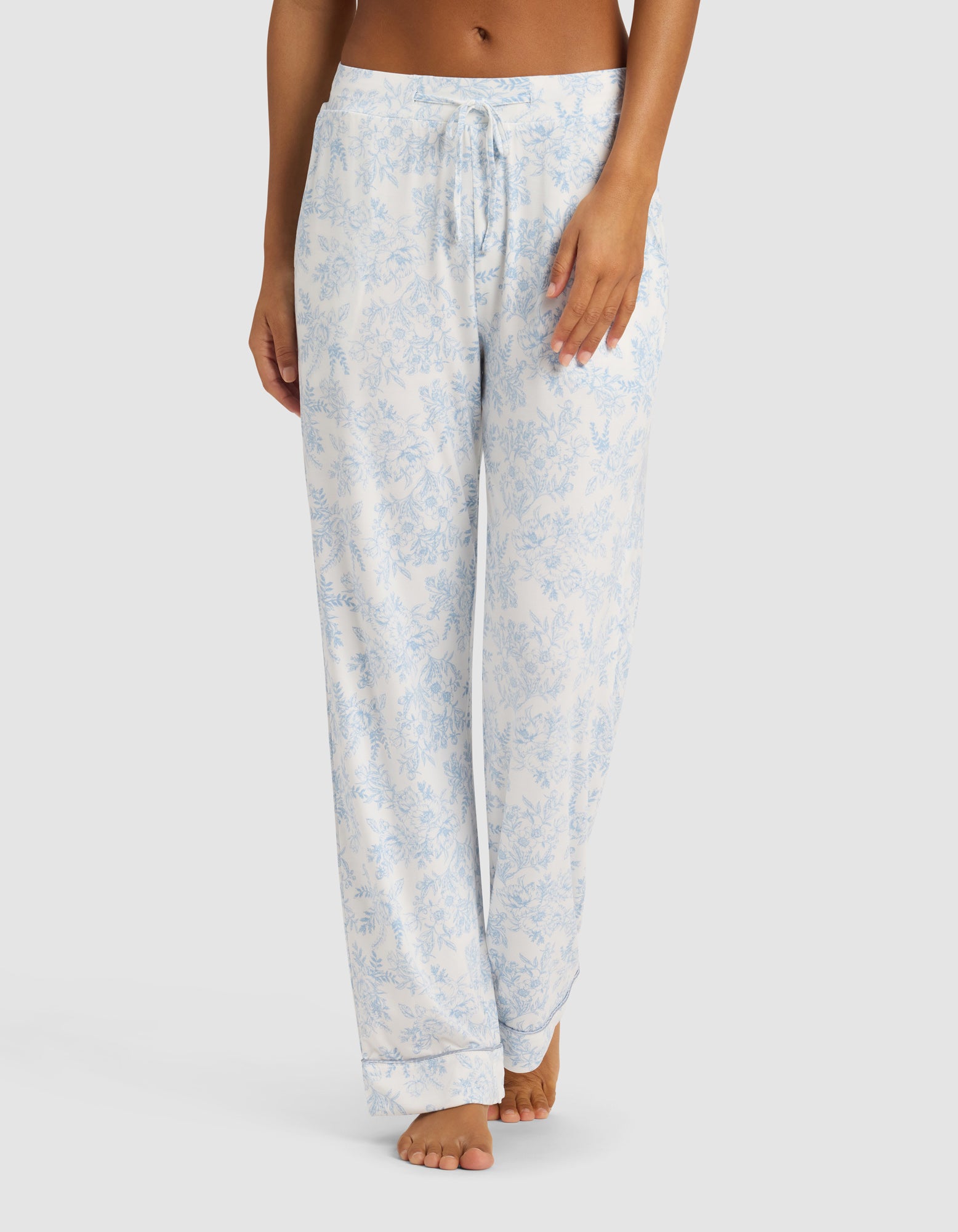 A woman wears Cozy Earth's Women's Bamboo Stretch-Knit Long Sleeve Pajama Set in light blue and white floral, featuring a drawstring waist, standing barefoot against a plain white background.