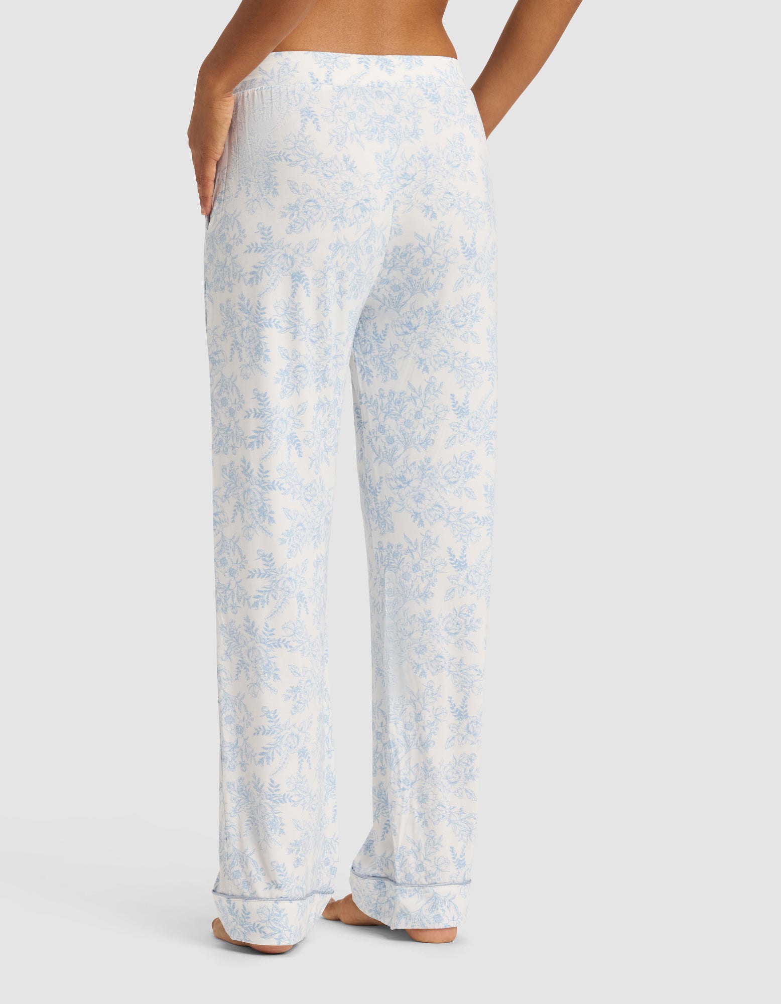 A person stands barefoot facing away, wearing Cozy Earth's Women's Bamboo Stretch-Knit Long Sleeve Pajama Set in white with a light blue floral pattern. Their right hand is on their hip against a plain light gray background.