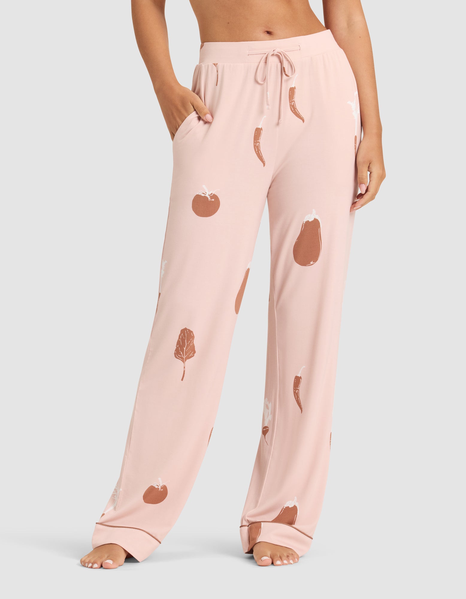 A person stands barefoot in HIDE's Women's Bamboo Stretch-Knit Pajama Pant—loose, light pink, with a drawstring waist, pockets, and brown vegetable prints featuring tomatoes, eggplants, peppers, and leafy greens. 