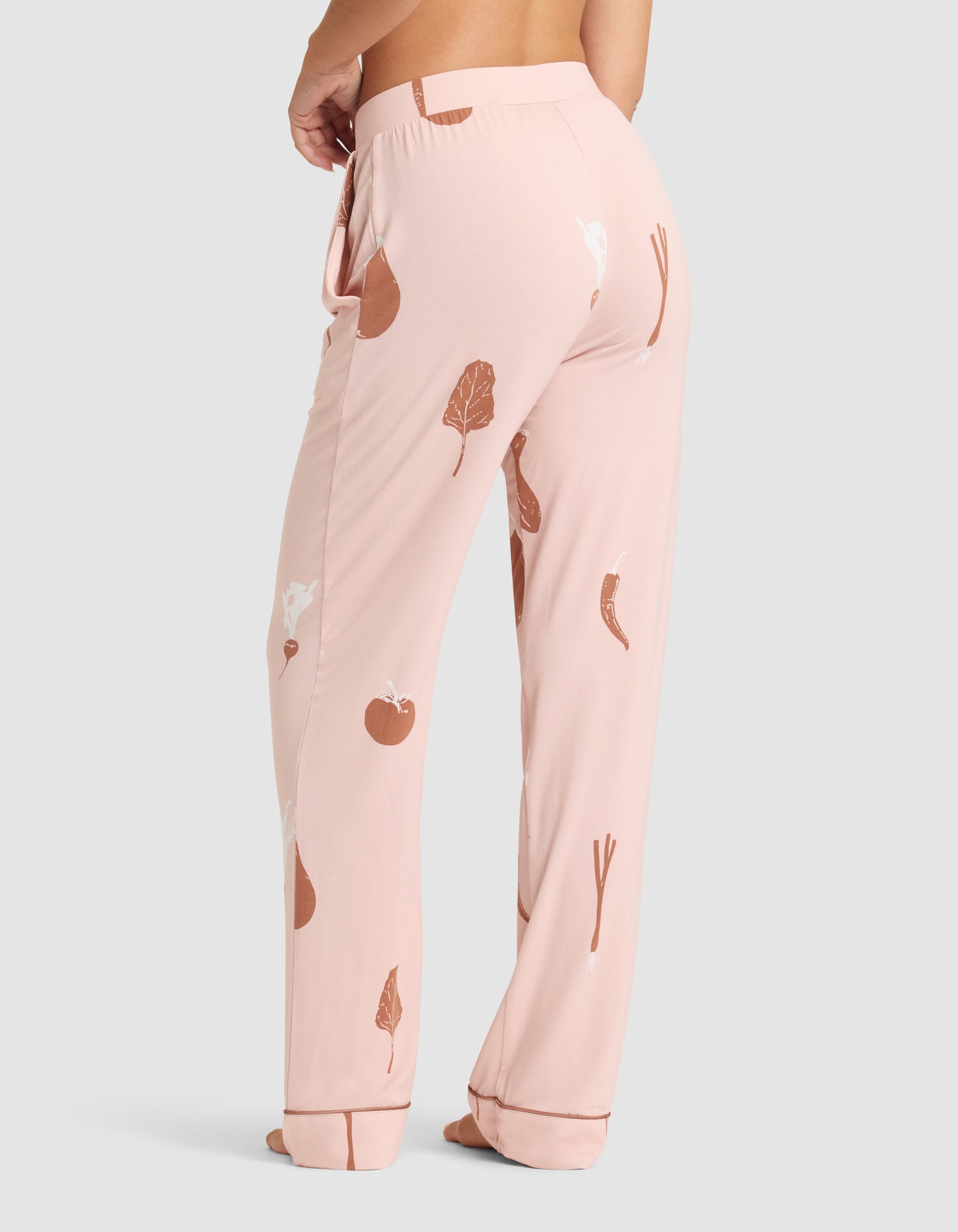 A person is barefoot wearing HIDE Women's Bamboo Stretch-Knit Pajama Pant in light pink with brown and white vegetable prints, shown from the lower back and legs against a plain light gray background. 