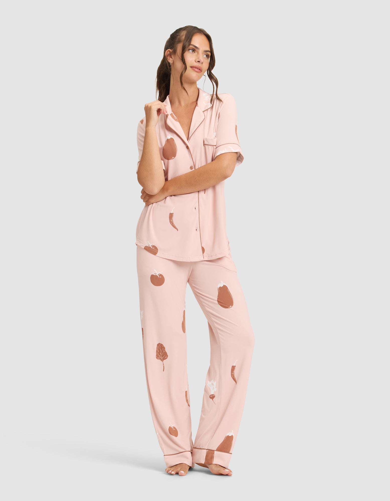 A woman smiles to the side with hands by her sides, wearing HIDE Women's Bamboo Stretch-Knit Pajama Pant—a light pink, loose fit set with a button-up short-sleeve top and pants, featuring brown and white abstract prints. She is barefoot. 