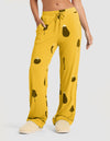 A person in HIDE Women's Bamboo Stretch-Knit Pajama Pant (Veggie), featuring mustard yellow fabric with black veggie prints and a drawstring waist, pairs them with cream slippers and stands with one hand in their pocket against a light background. |Color:Veggies in Mango