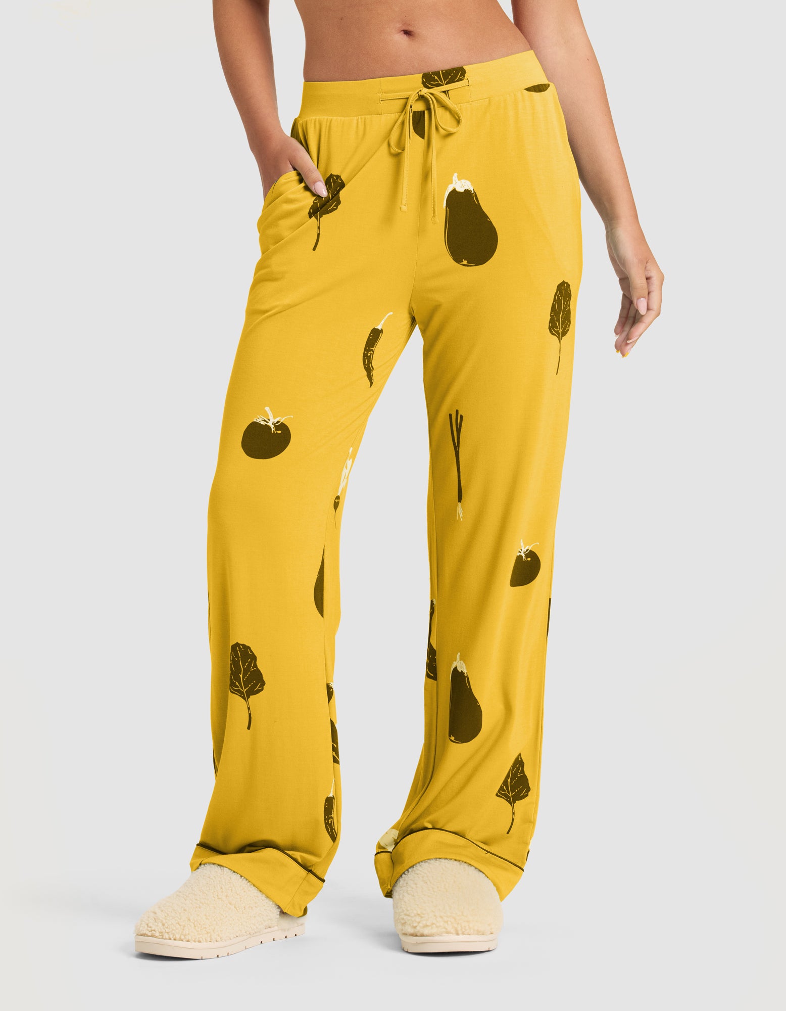 A woman wears Cozy Earth's Women's Bamboo Stretch-Knit Short Sleeve & Pant Pajama Set (Veggie), featuring loose-fitting yellow veggie-print pants with pockets and a drawstring waist, paired with cream slippers against a plain light gray background. 