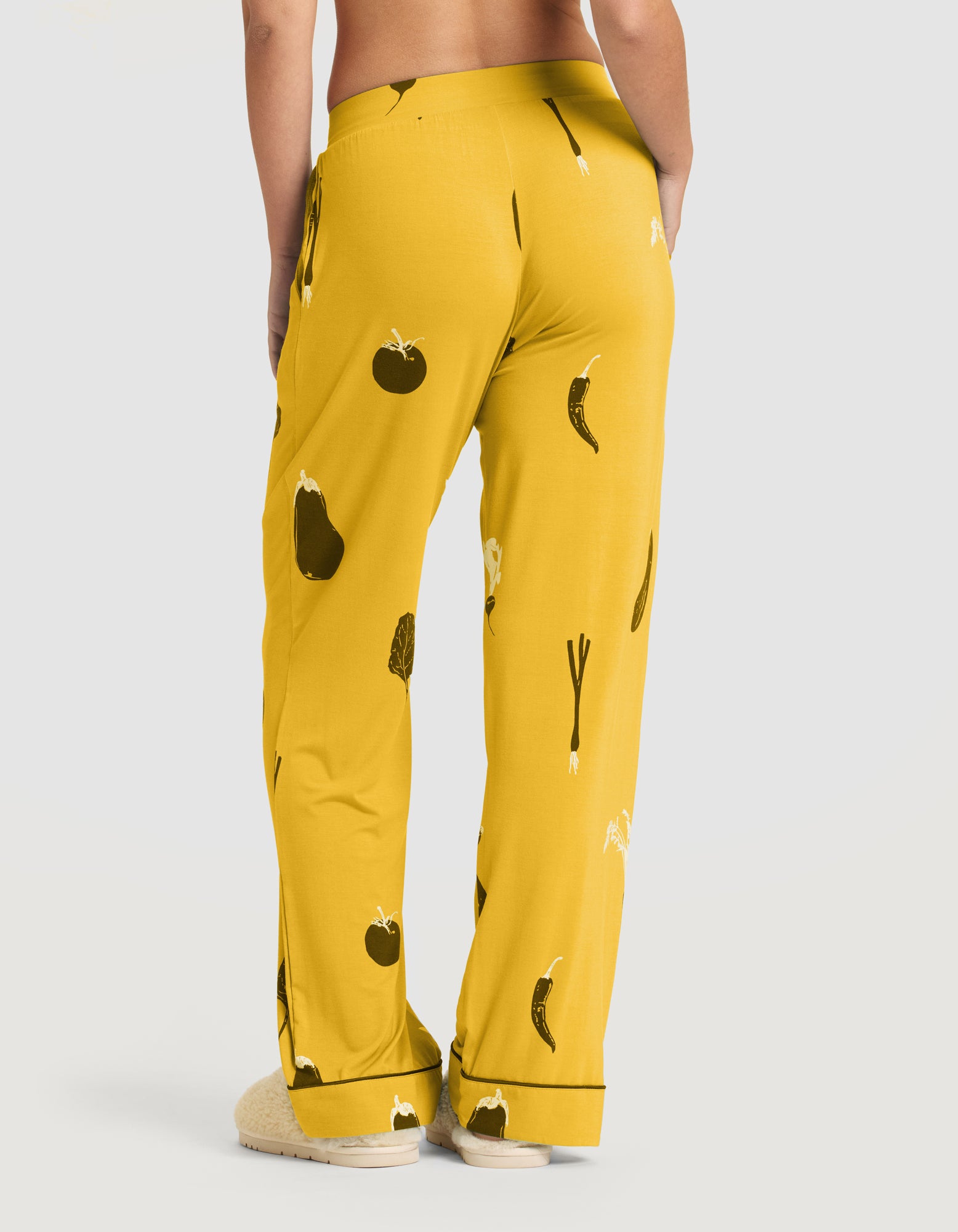 A person shown from the back wears HIDE's Women's Bamboo Stretch-Knit Pajama Pant (Veggie), featuring a yellow veggie print, paired with cream fuzzy slippers. 